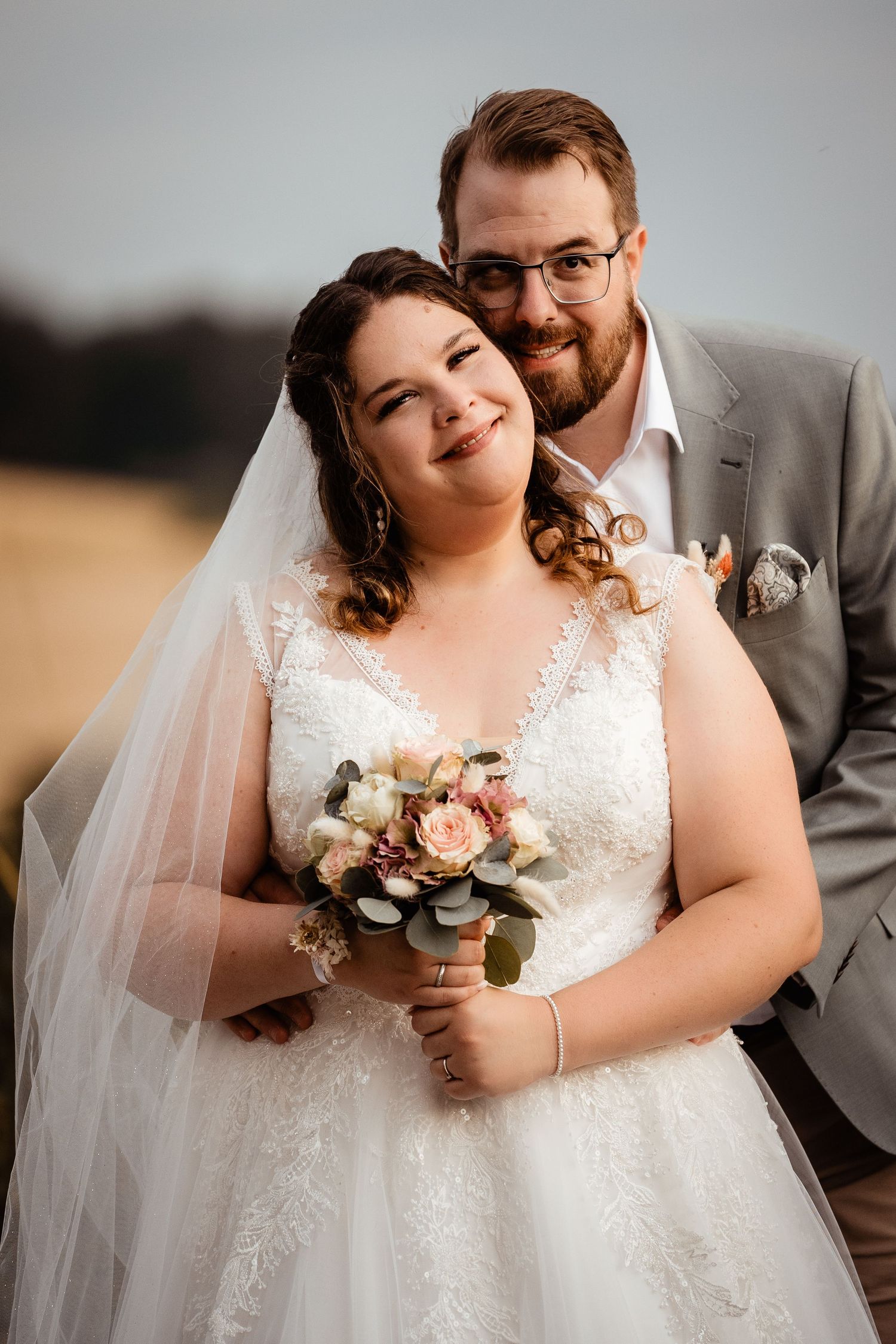 Bride and groom pose at golden hour, she holds a pastel bouquet, he wears a gray suit, field background.