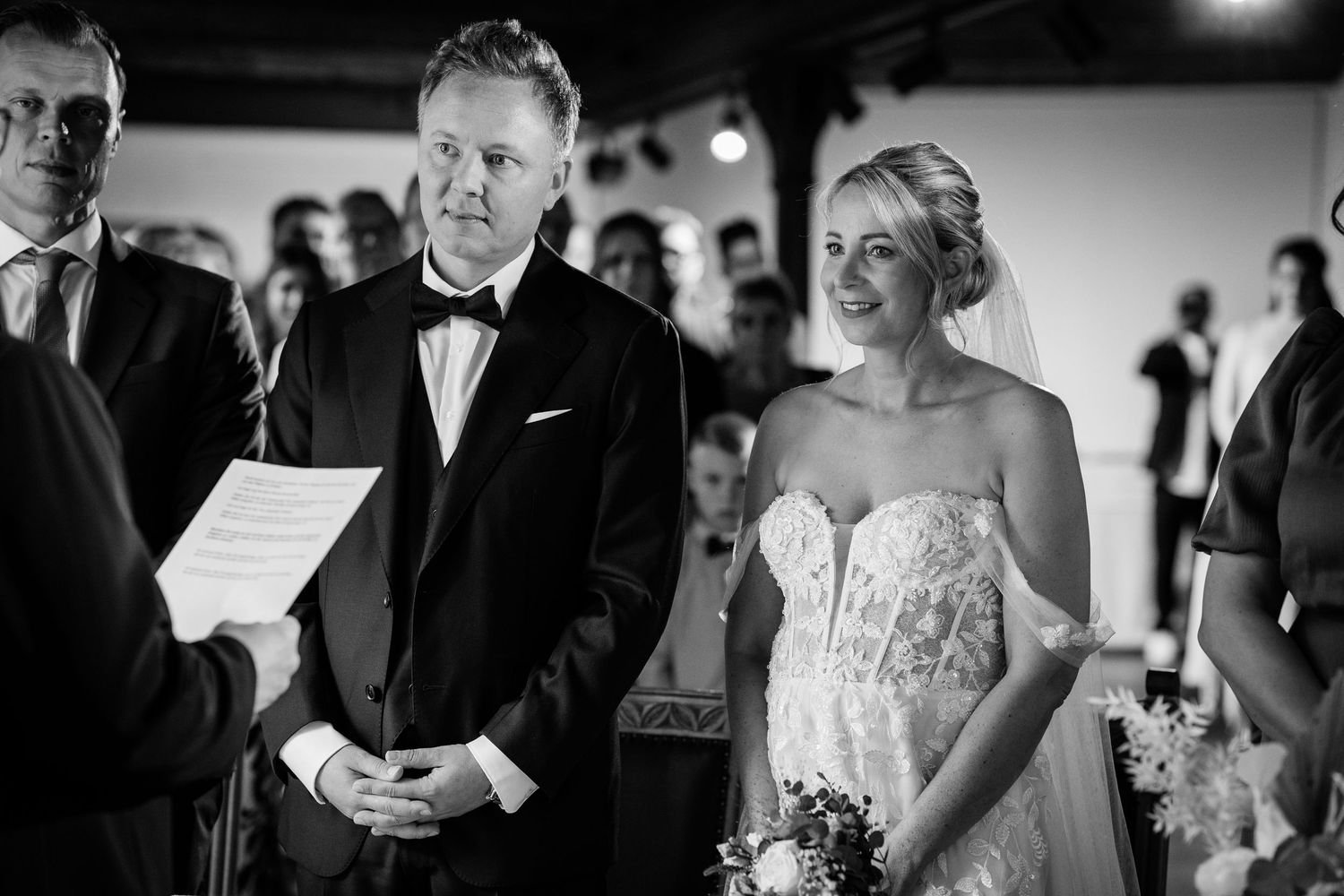 Black and white photo of a smiling bride in lace gown and groom in tuxedo during wedding ceremony.