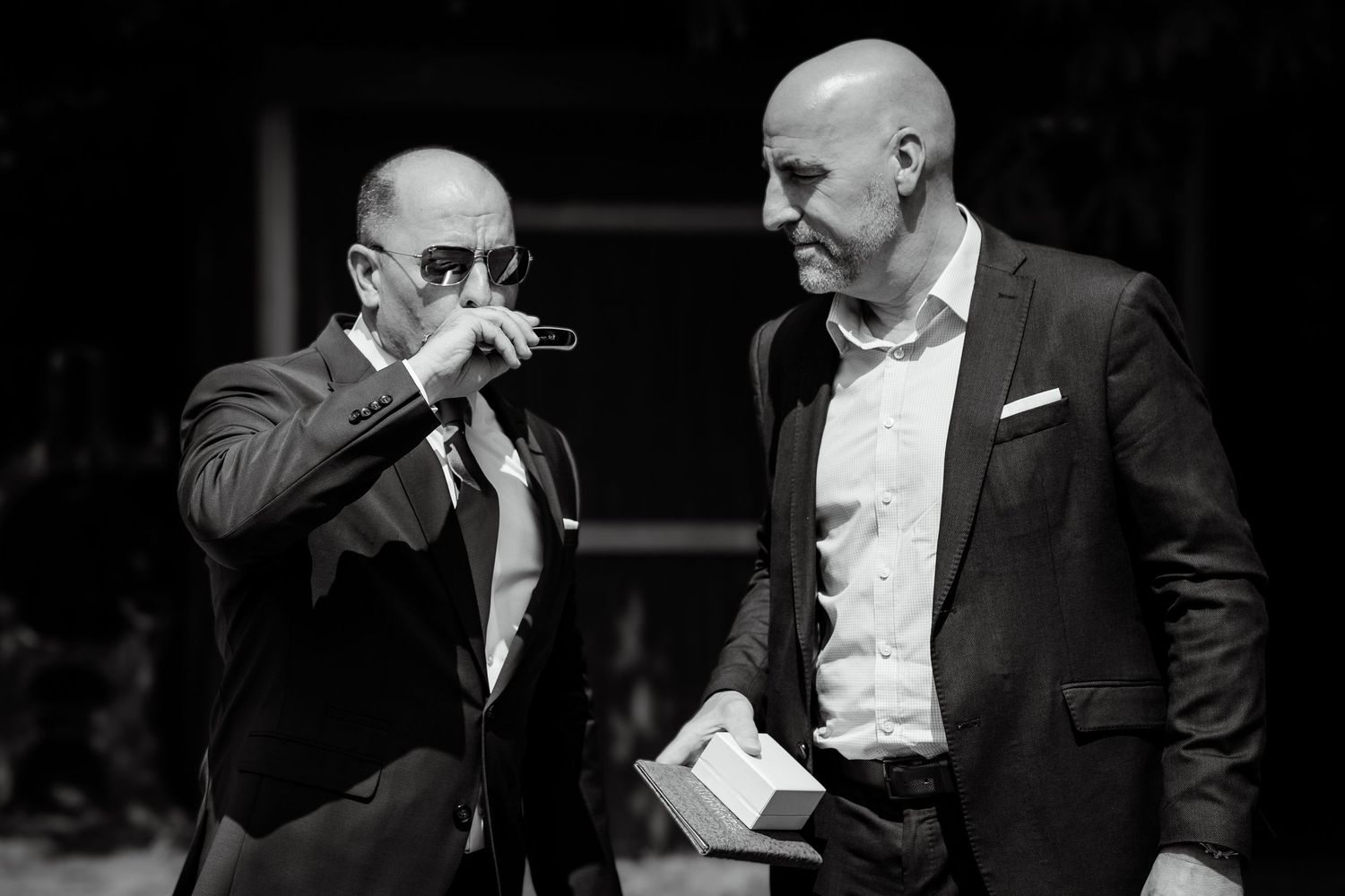 Two men in suits conversing at a formal event, one adjusting glasses, black and white photography.