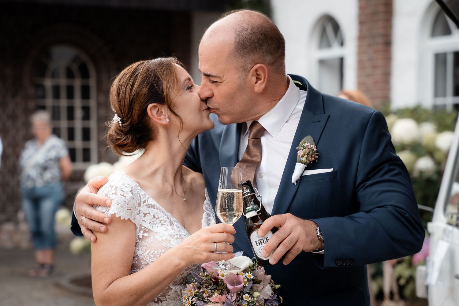 Bride and groom share a kiss while holding champagne glasses outdoors after their wedding ceremony.
