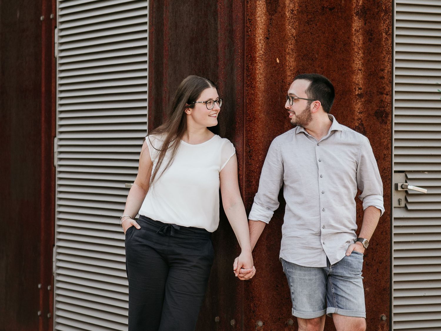 A couple in casual attire holds hands while walking past a rustic corrugated metal wall and wooden beams.