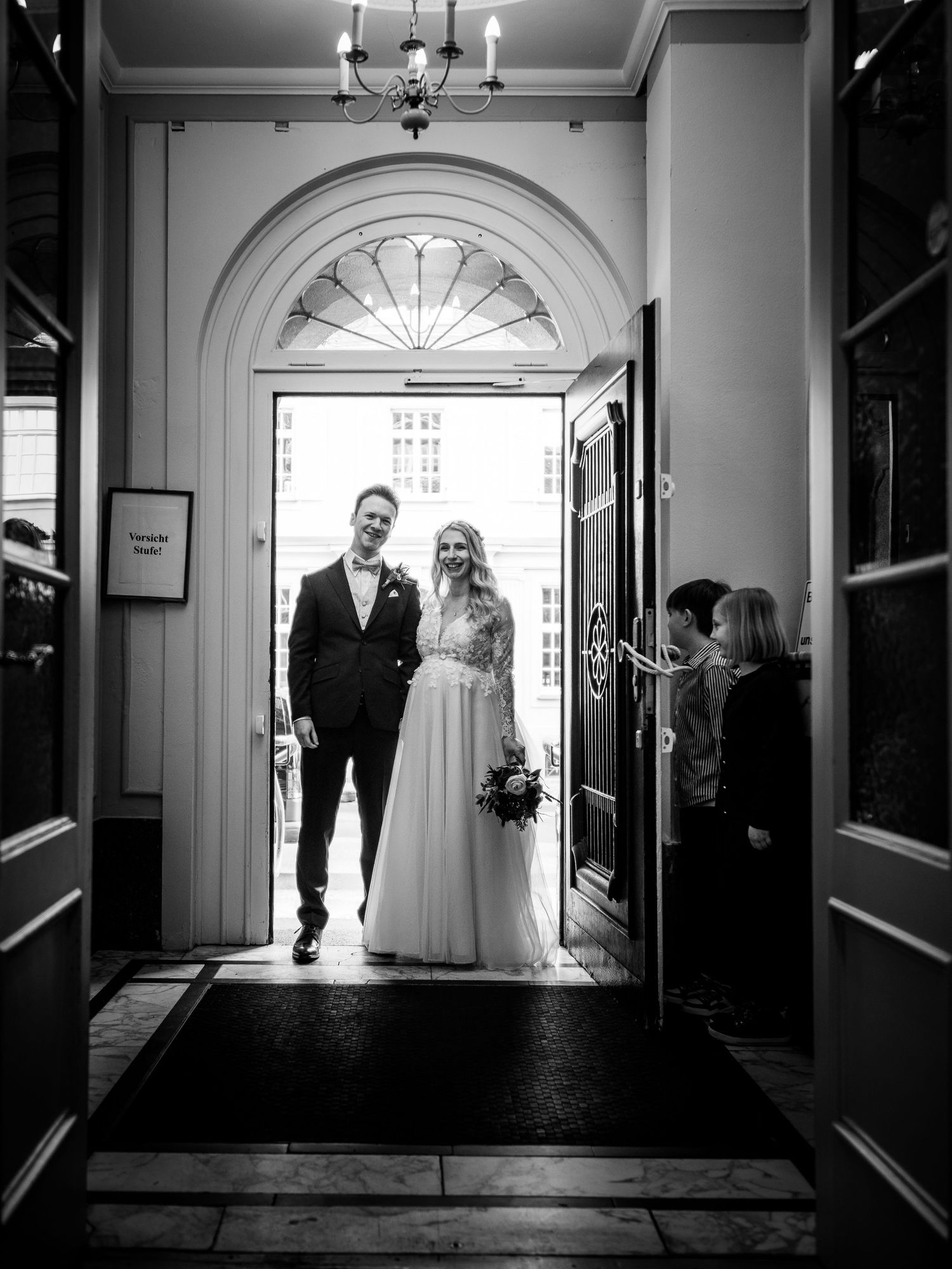 Bride and groom smiling as they exit through an elegant arched doorway in a black and white wedding photo.