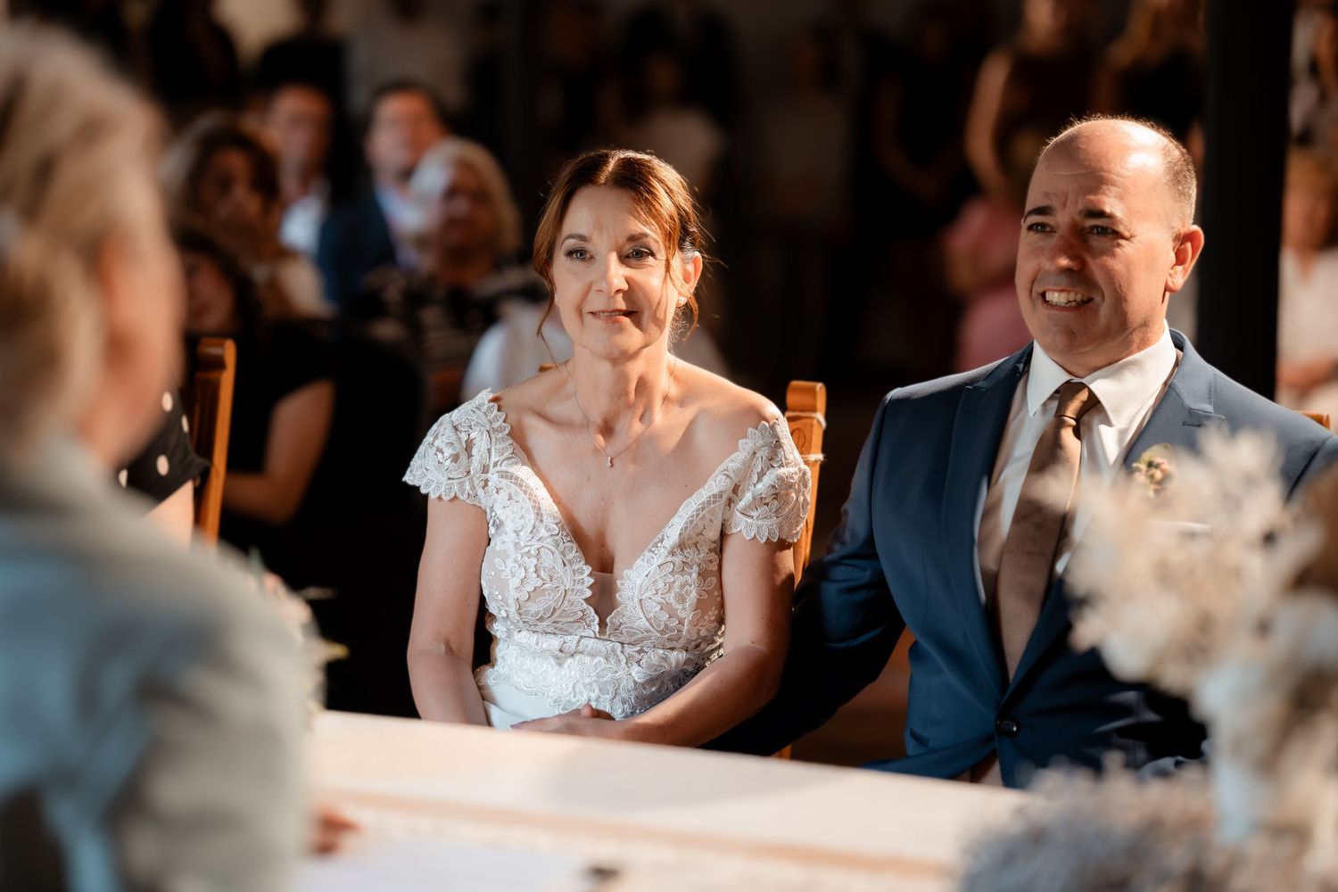 Bride and groom smiling joyfully during wedding ceremony, seated at decorated table with floral arrangements.