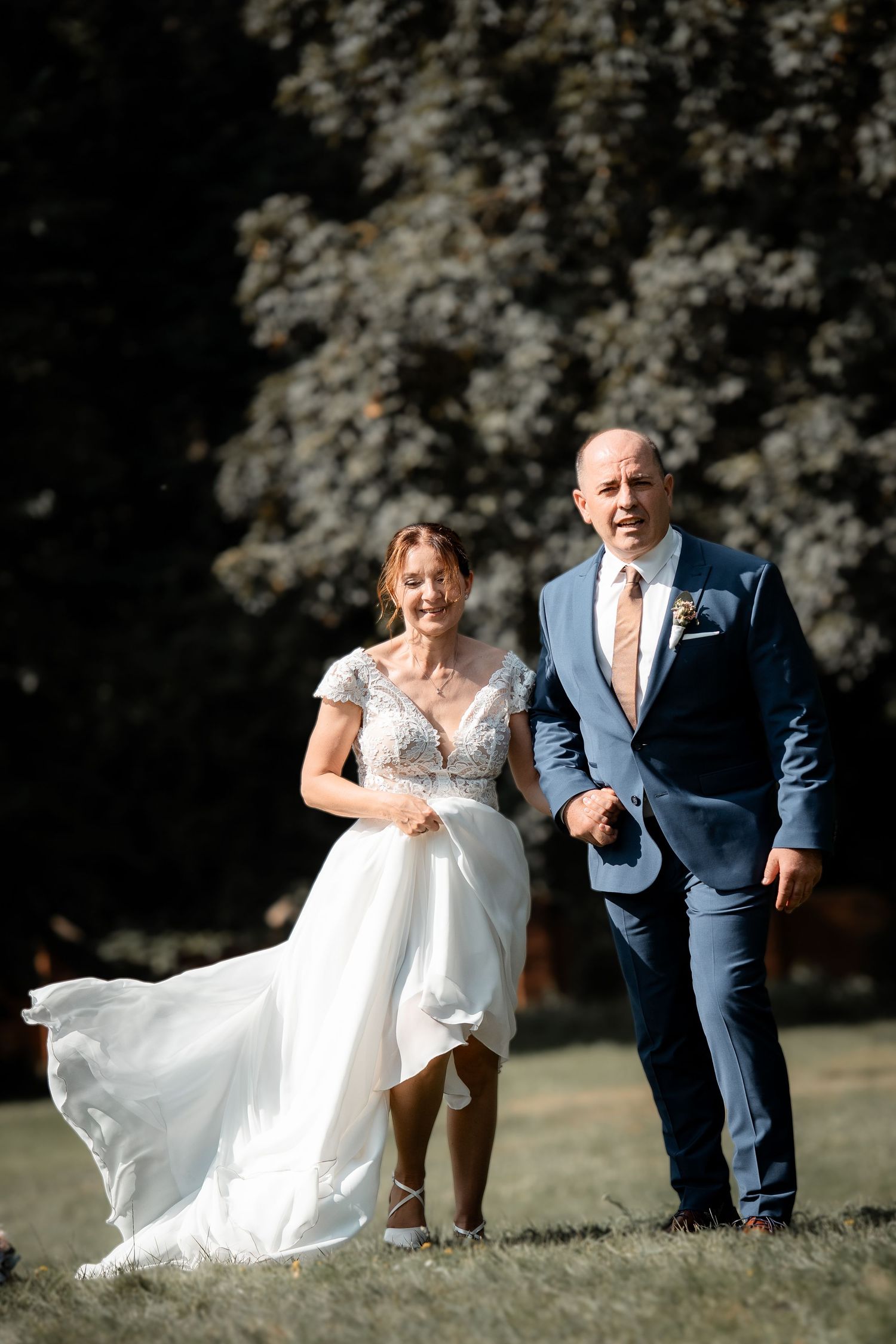 Bride in white lace gown and groom in blue suit walk together across sunny outdoor garden lawn.