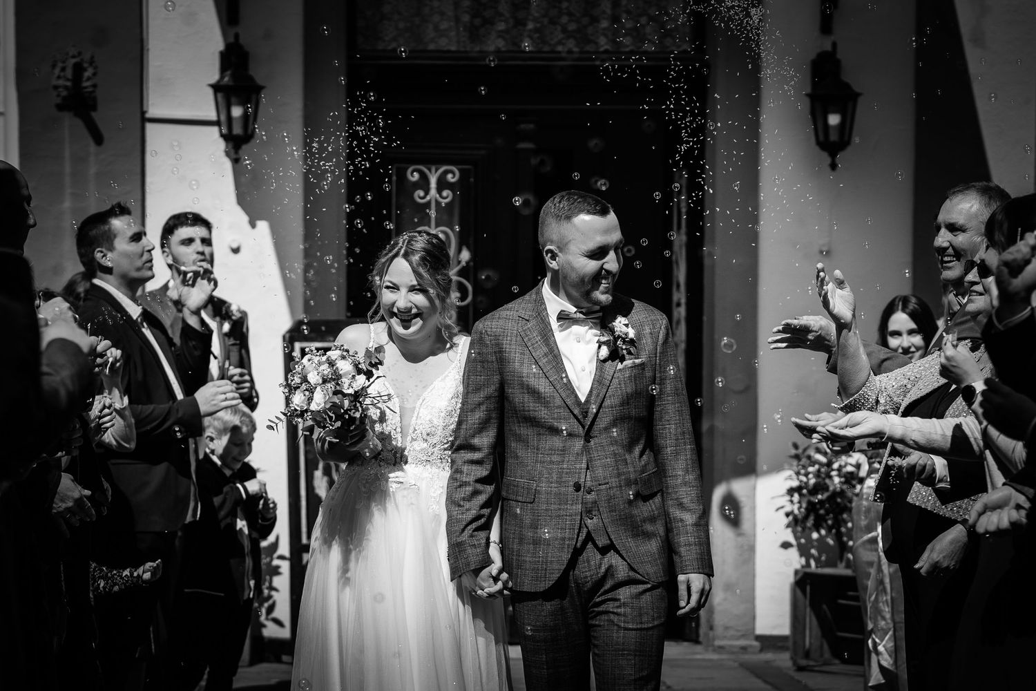 Bride and groom exit church amid confetti shower, surrounded by smiling guests in black and white wedding photo.