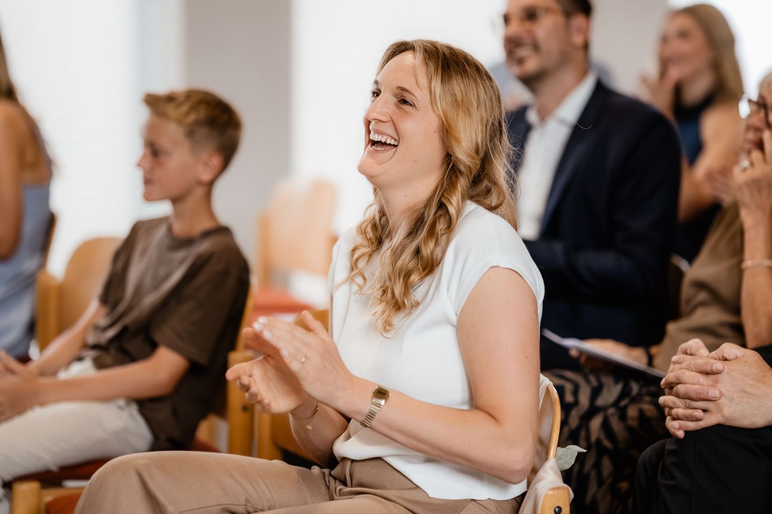 Happy woman laughing and clapping while seated at an indoor event surrounded by other attendees.