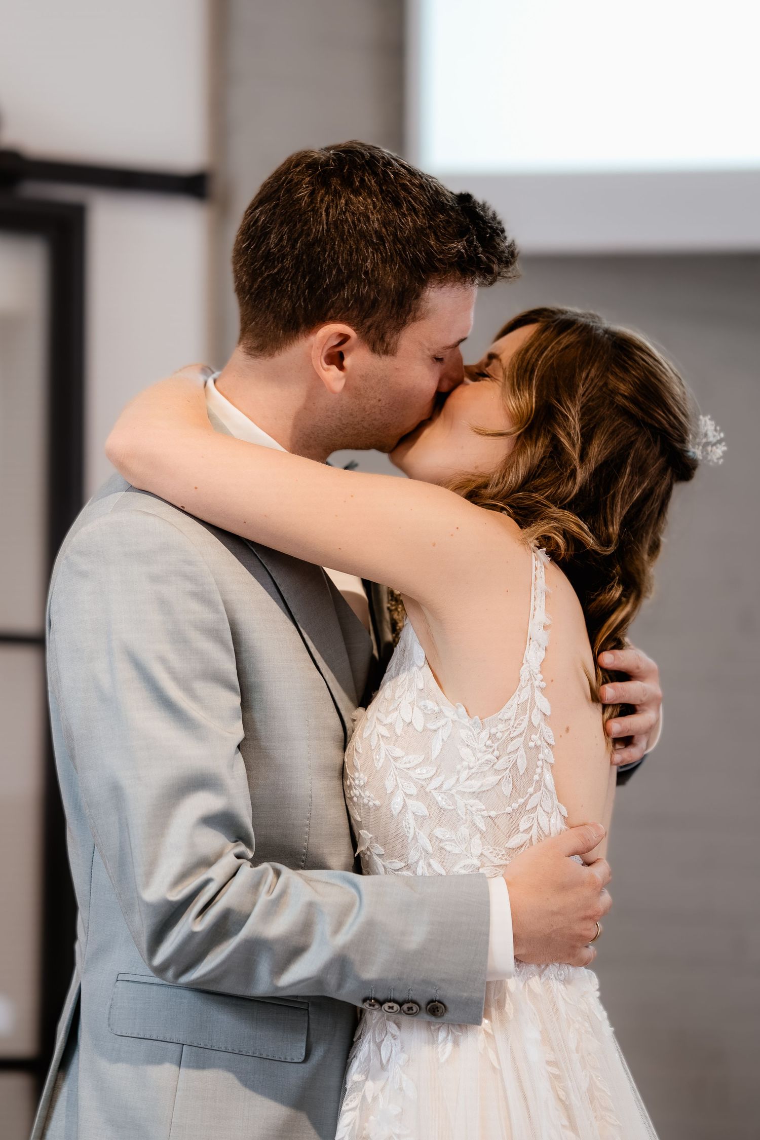 Bride and groom sharing first kiss, bride in lace wedding dress, groom in gray suit.