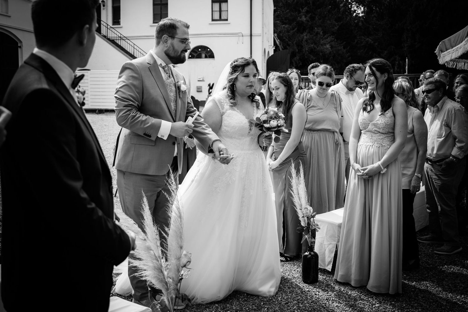 Black and white photo of bride in white gown and groom in gray suit greeting guests at outdoor wedding ceremony.