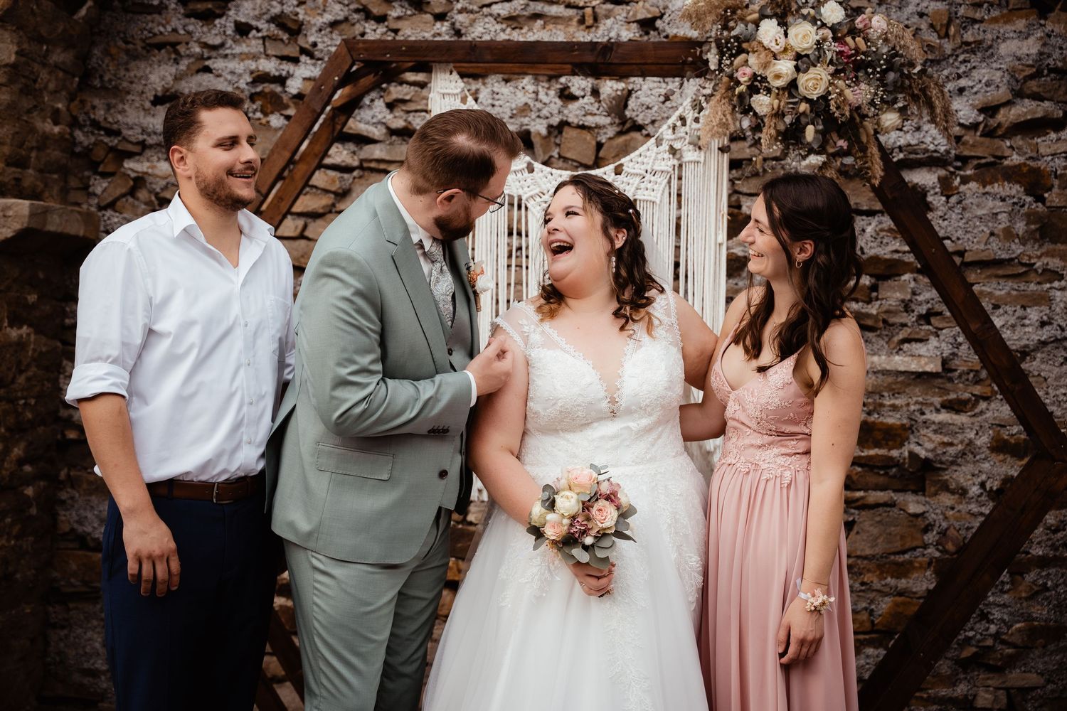 Laughing bride and groom in wedding attire pose with guests before a rustic stone wall and floral arch.