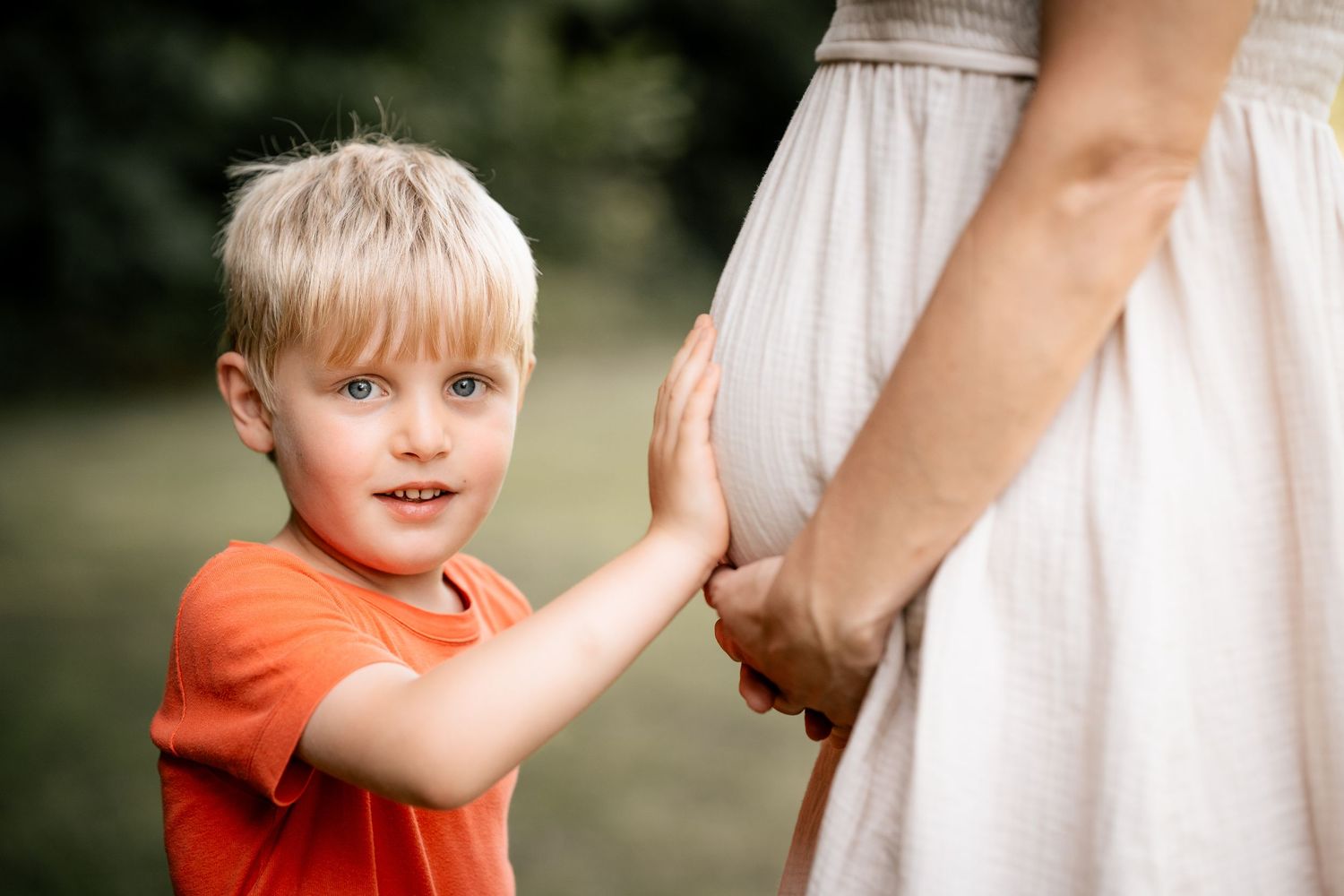 Blonde toddler boy in orange shirt touching pregnant mother's belly outdoors in a green park setting.