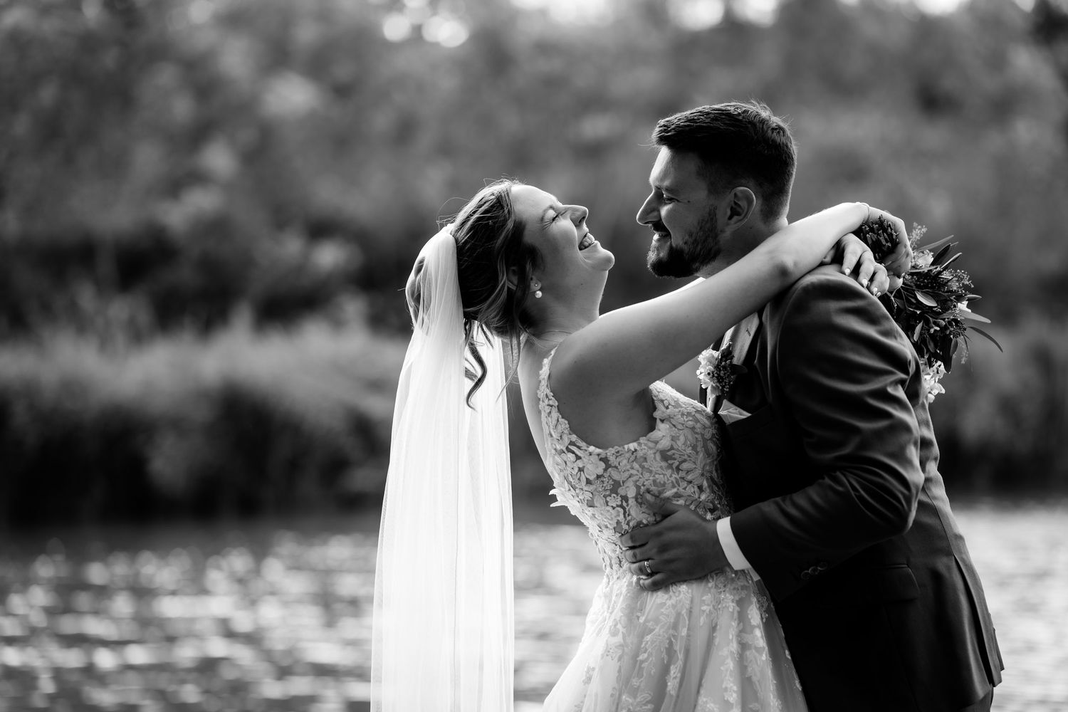 Black and white photo of a bride and groom sharing a romantic embrace near a serene lake on their wedding day.