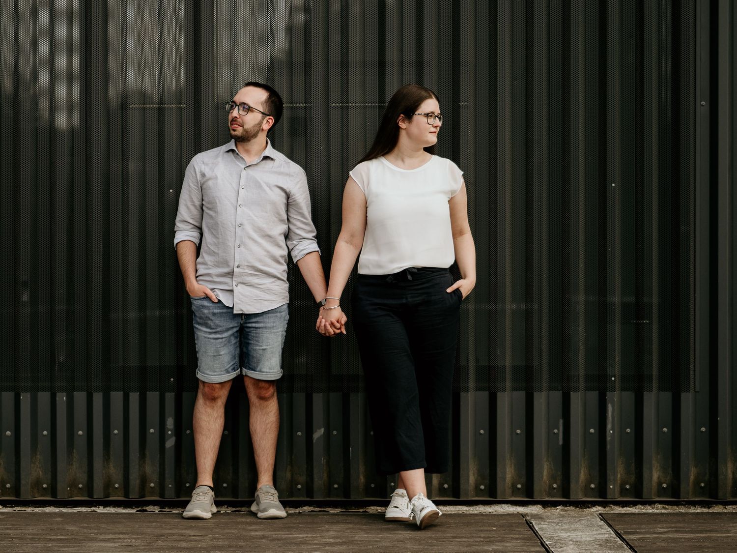 Couple holding hands standing against a dark corrugated wall, man in shorts and woman in black pants.