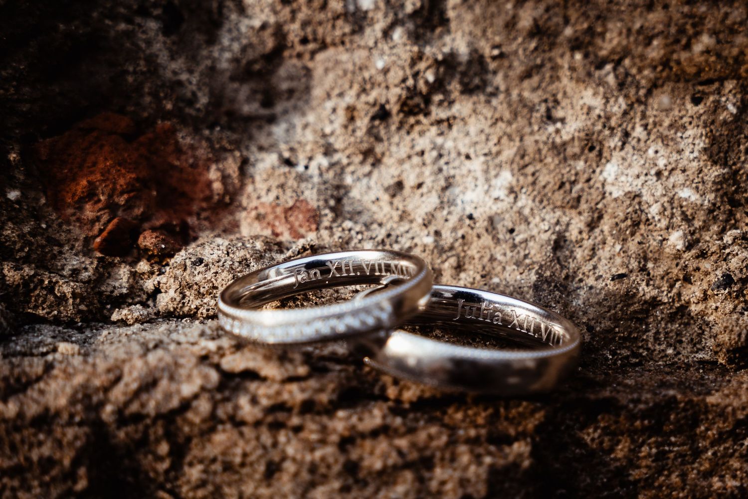 Two silver wedding bands with engraved Roman numerals resting on a textured stone surface.