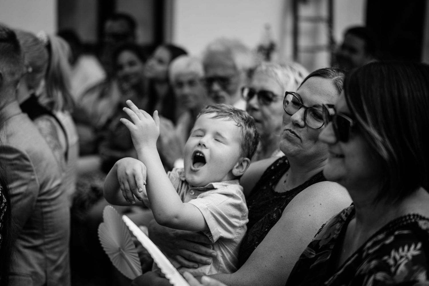 Black and white photo of a yawning toddler being held by a woman wearing sunglasses at a crowded outdoor event.