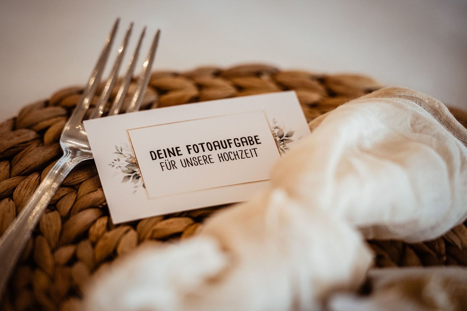 A wedding place card reading Deine Fotoaufgabe fur unsere Hochzeit rests beside a fork on a woven charger plate.