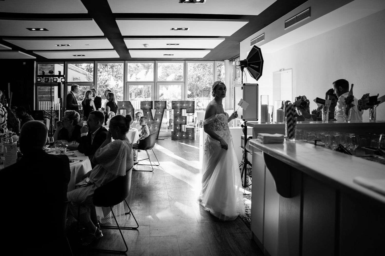 Black and white photo of a bride standing in a sunlit wedding reception venue with seated guests.