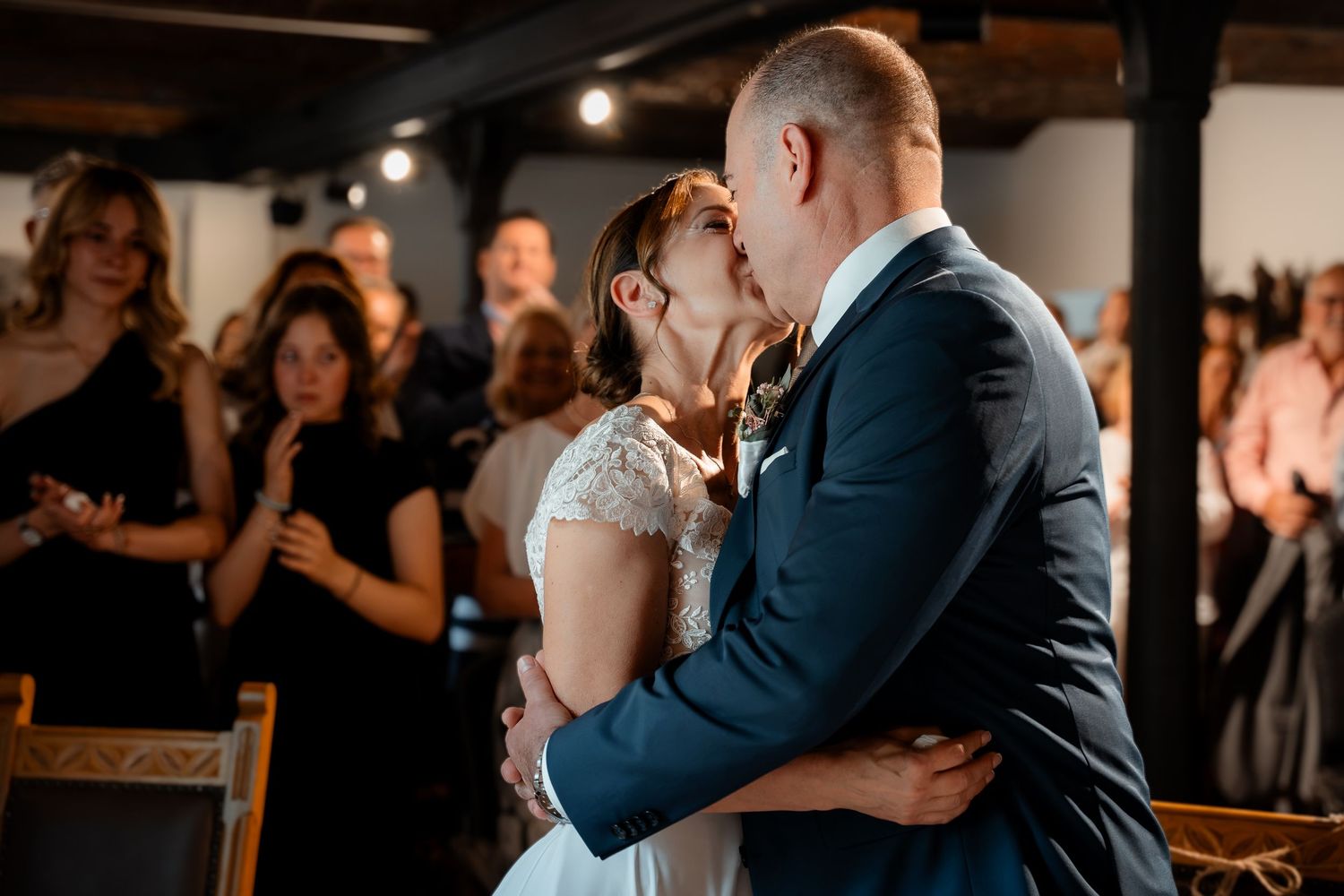 Bride and groom share first kiss during indoor wedding ceremony while guests applaud in background.
