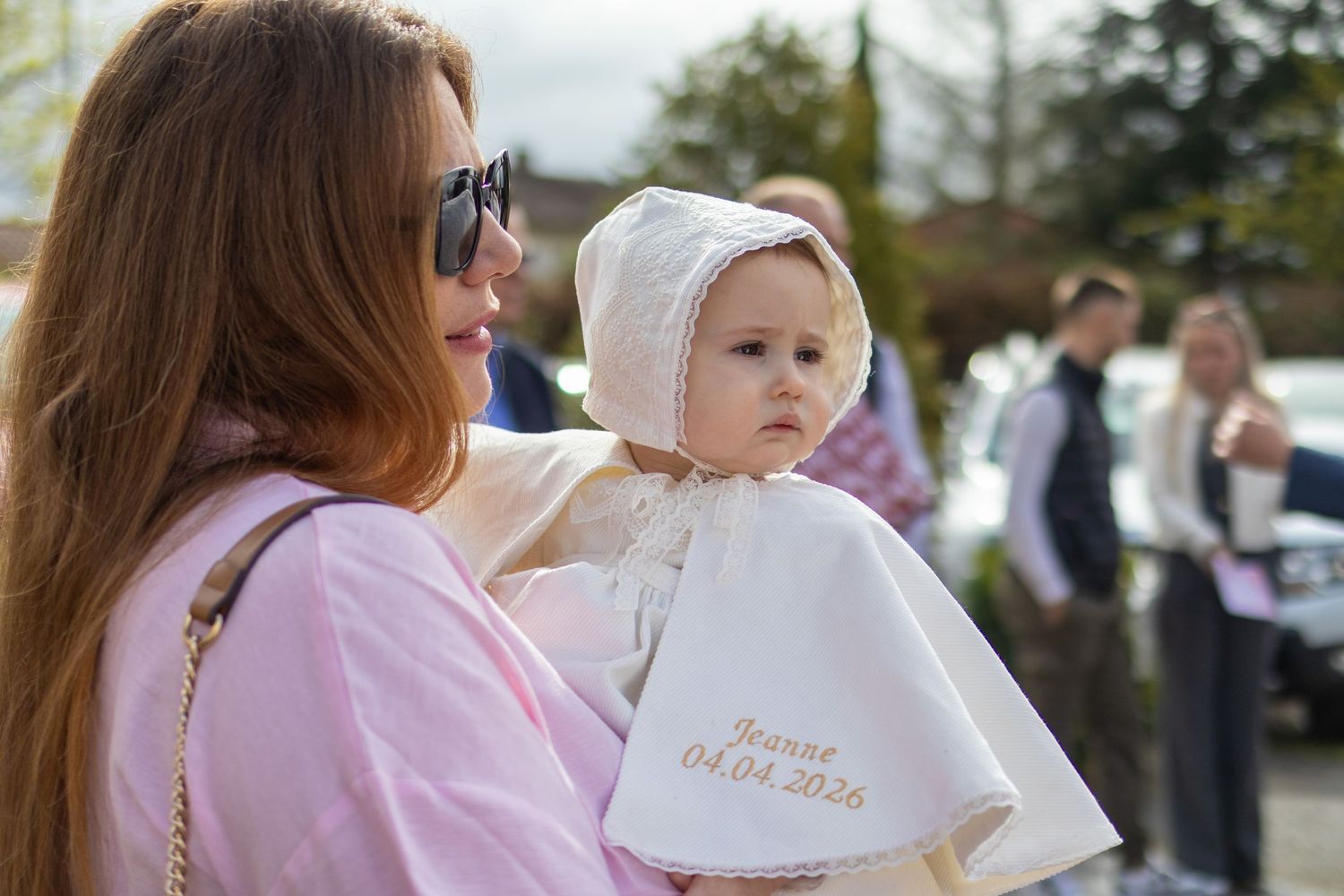 A mother in pink holds her baby dressed in a white christening gown and bonnet outdoors on a sunny day.