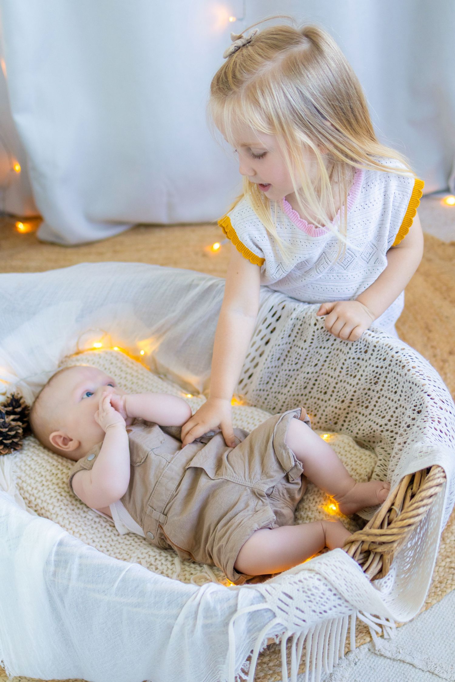 Blonde toddler girl lovingly watching over newborn baby sibling lying in a cozy wicker bassinet with warm fairy lights.