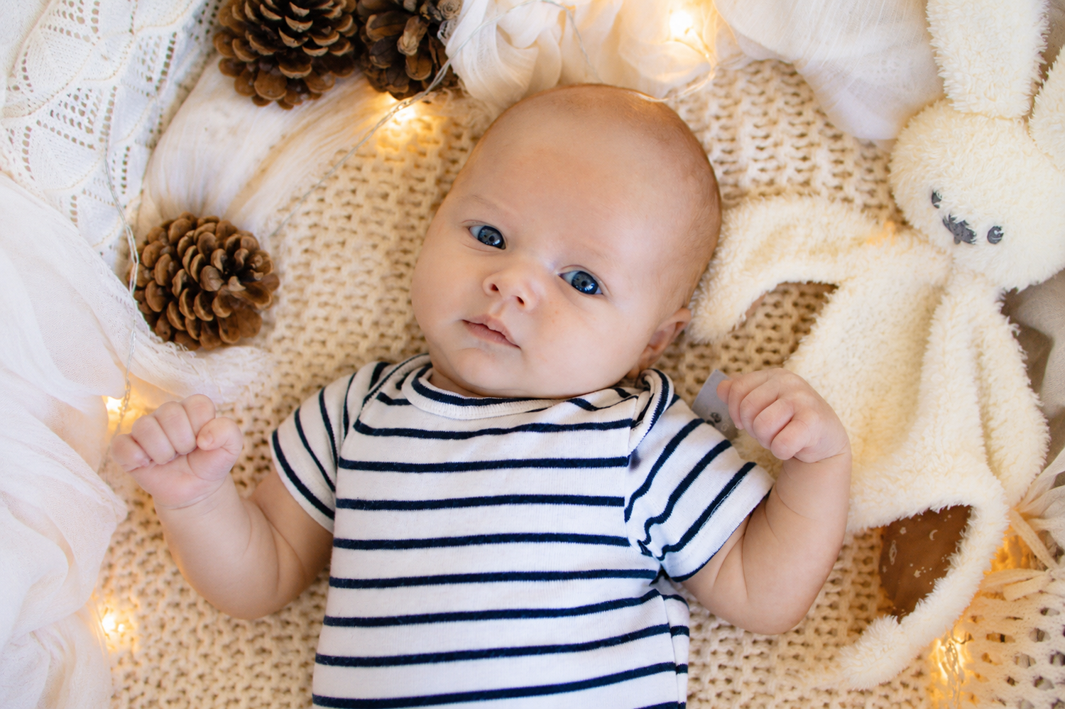 Blue-eyed baby in striped onesie lying on knit blanket with pine cones, fairy lights, and stuffed bunny toy.