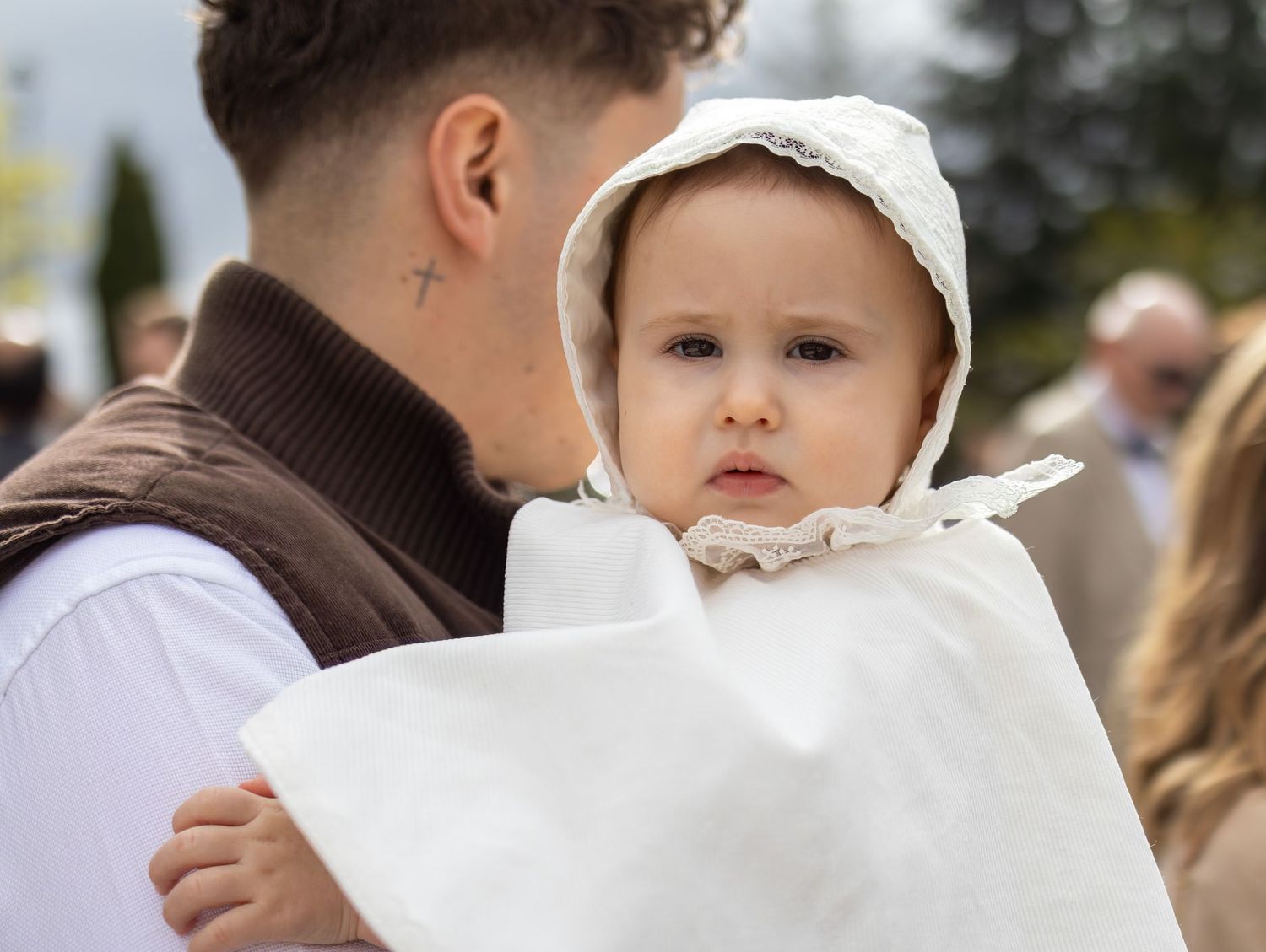 A baby in white christening gown and bonnet being held by a young man with a cross tattoo on his neck.
