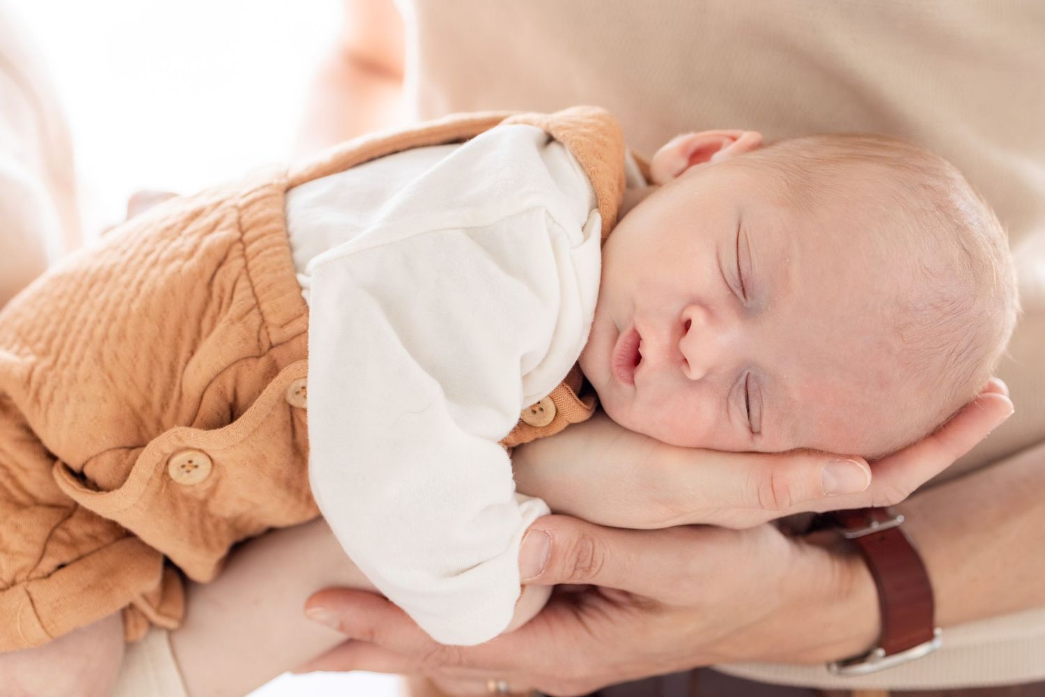 Sleeping newborn baby cradled in adult hands, wearing a white onesie and tan outfit.