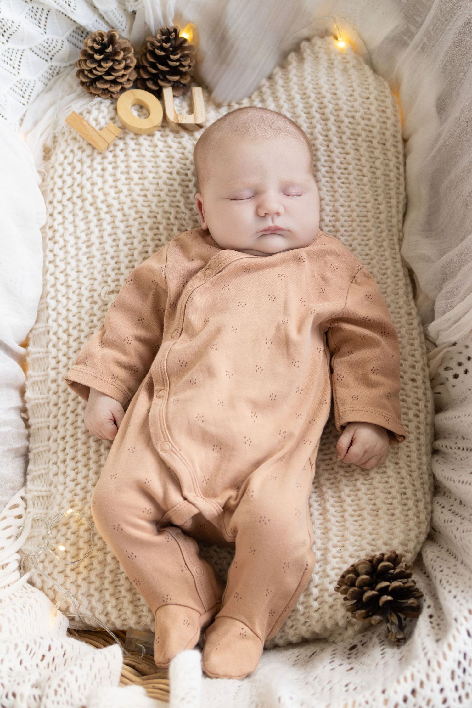 Newborn baby sleeping peacefully in a brown onesie on a knit pillow with pine cones and festive decorations.