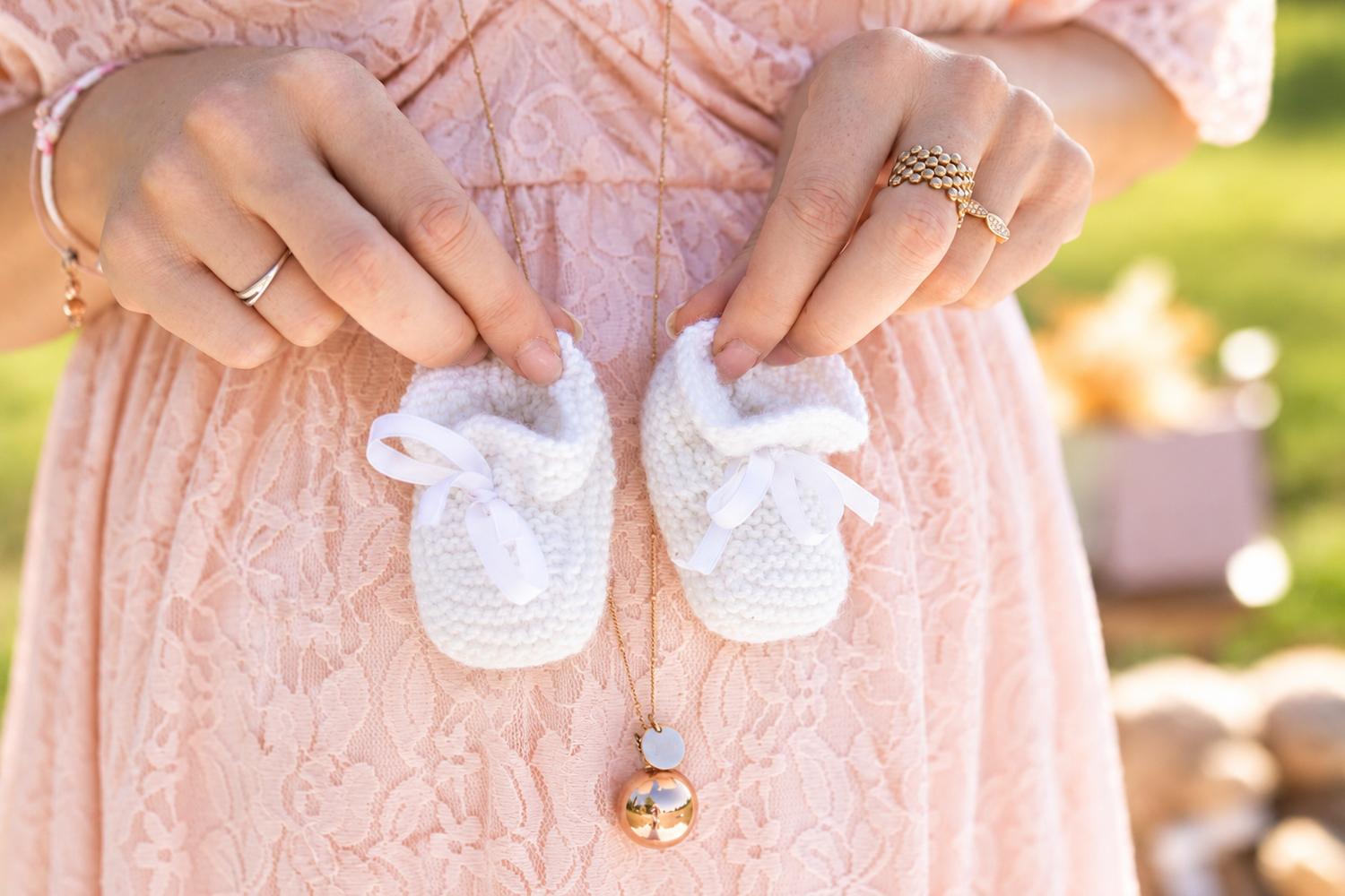 Pregnant woman in pink lace dress holding tiny white knitted baby booties over her baby bump.
