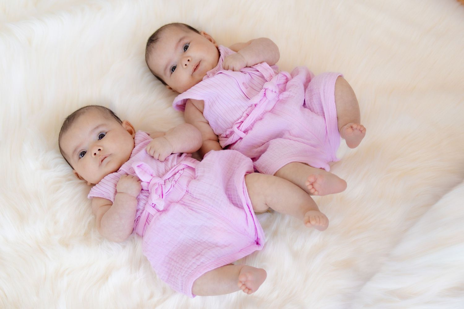 Two adorable twin baby girls in matching pink outfits lying on a soft white fluffy blanket, smiling cutely.
