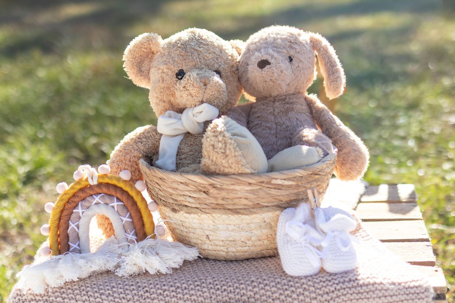 Two cute teddy bears sitting together in a wicker basket with a rainbow decoration and white booties outdoors.