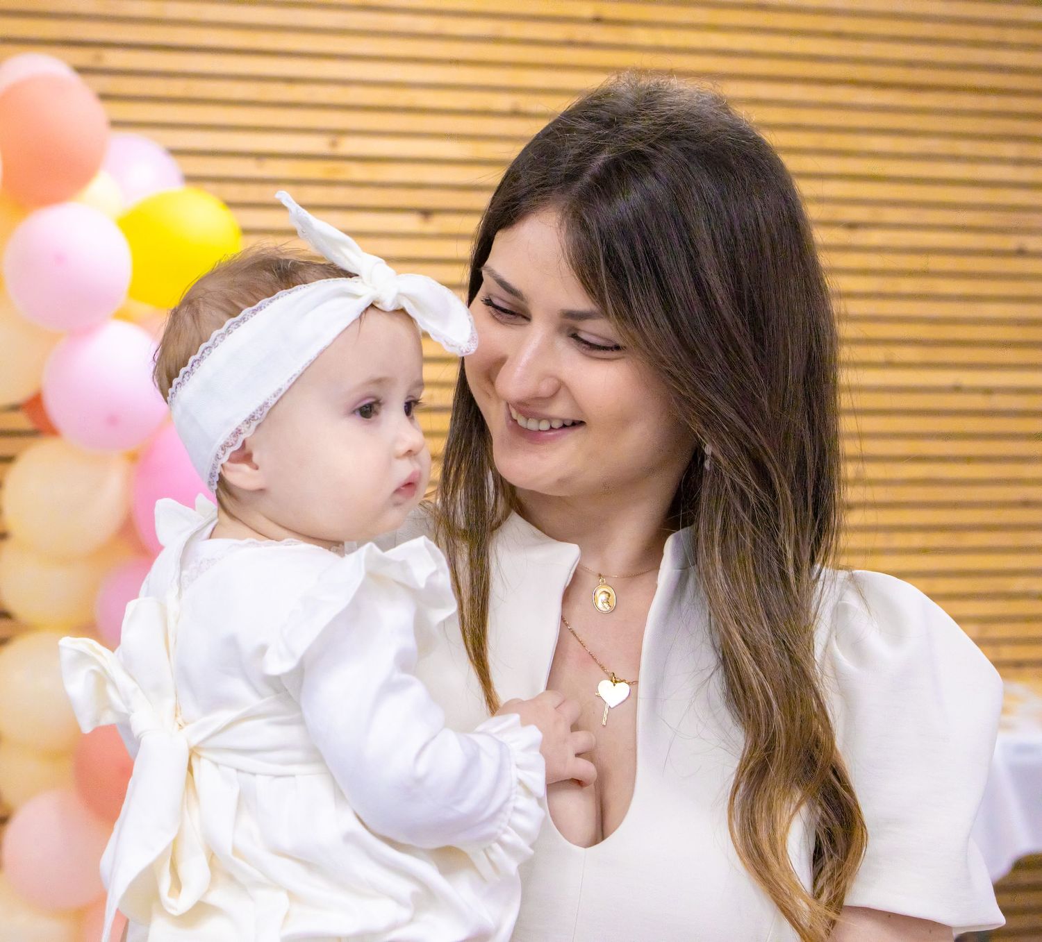 A smiling woman in white holds a baby girl in a white dress with a bow headband at a pink balloon party.