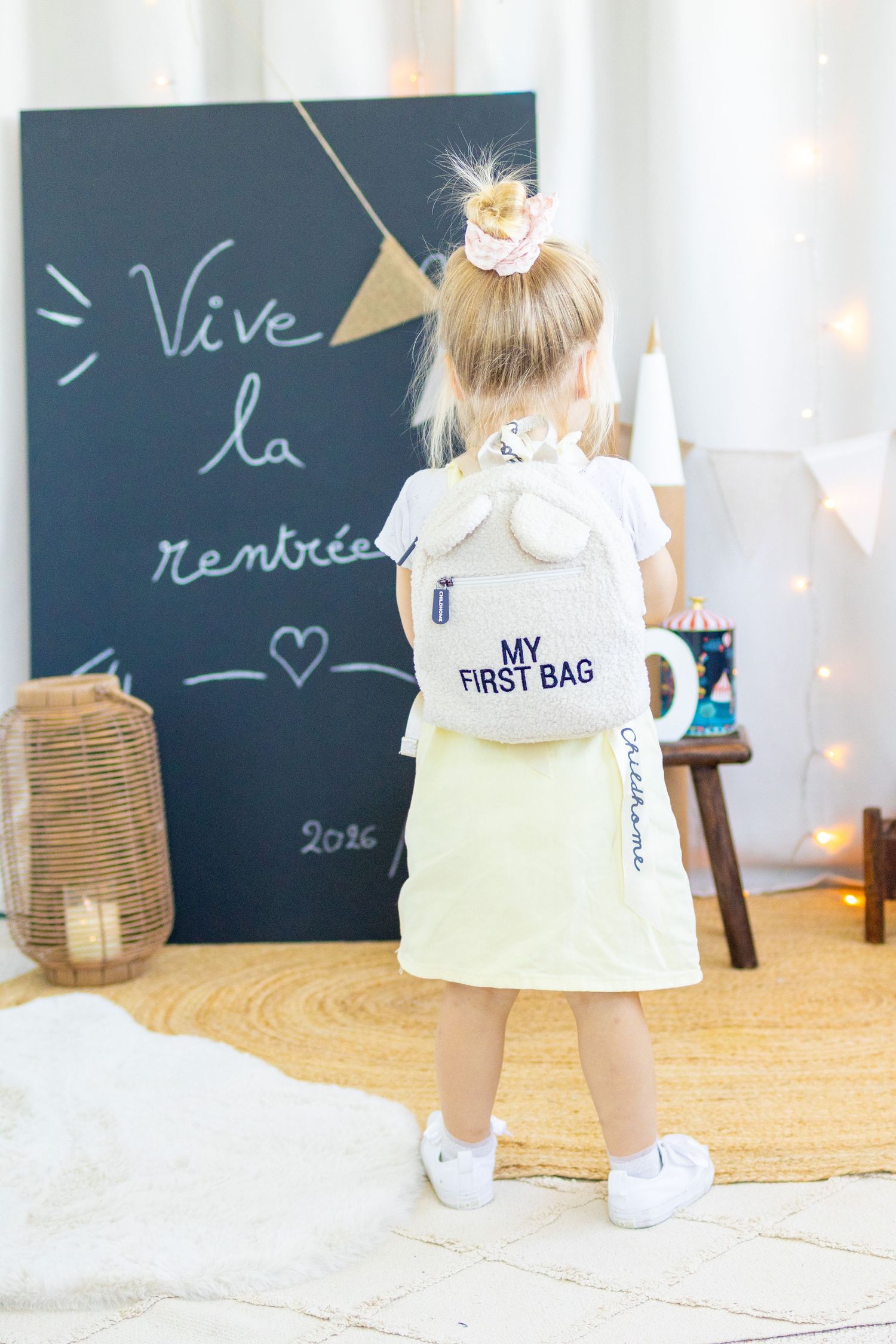 Young girl wearing a white bunny ear backpack labeled My First Bag, standing before a chalkboard reading Vive la rentree.