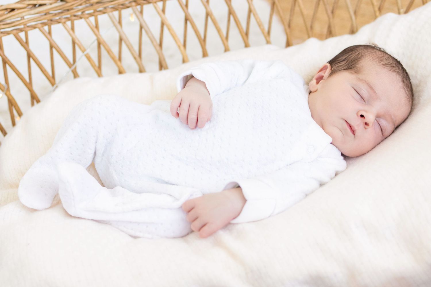 Peaceful newborn baby sleeping in a white onesie on a soft fur blanket inside a wicker bassinet.