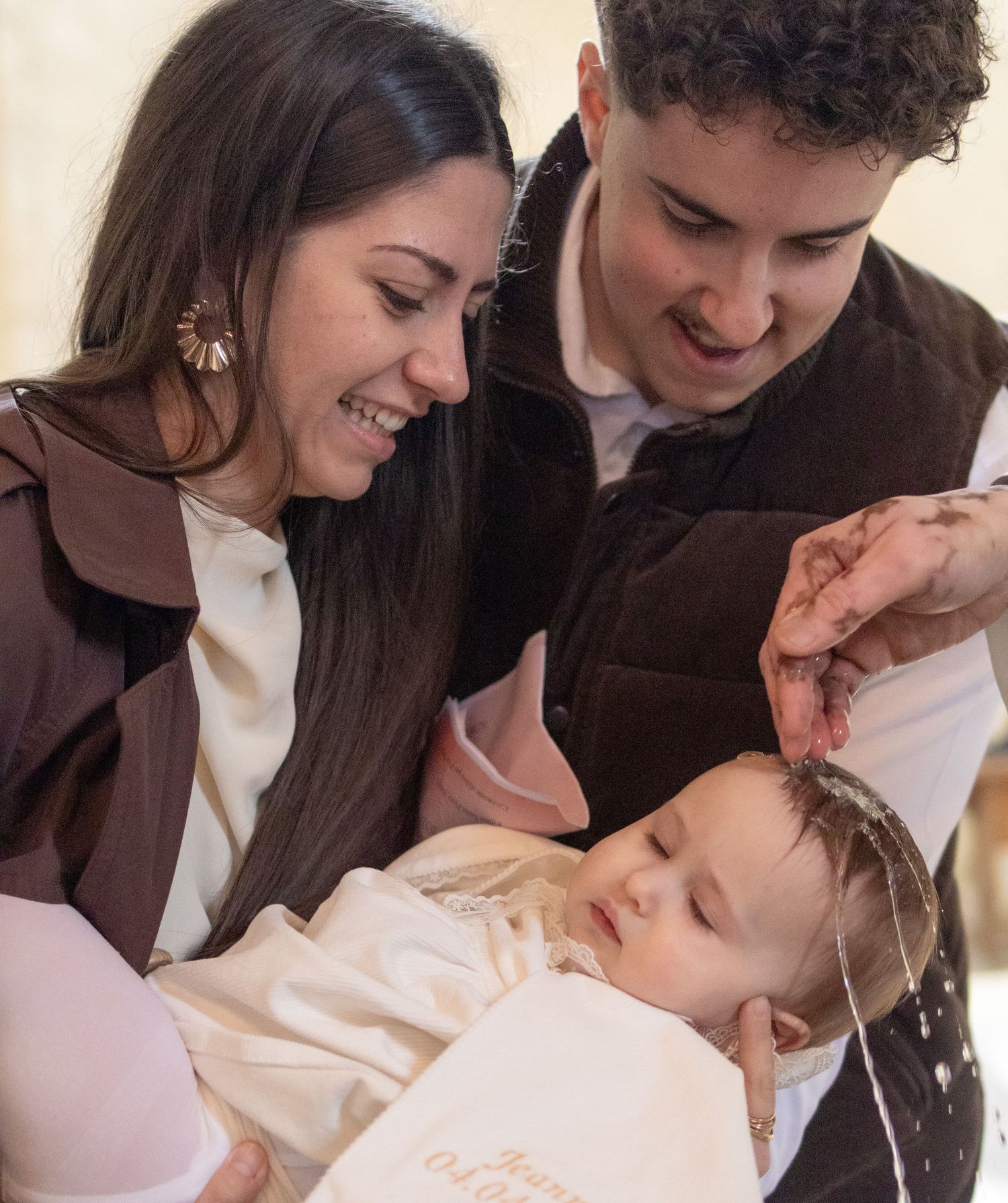 A baby being baptized with water poured on head, held by smiling young parents during a christening ceremony.