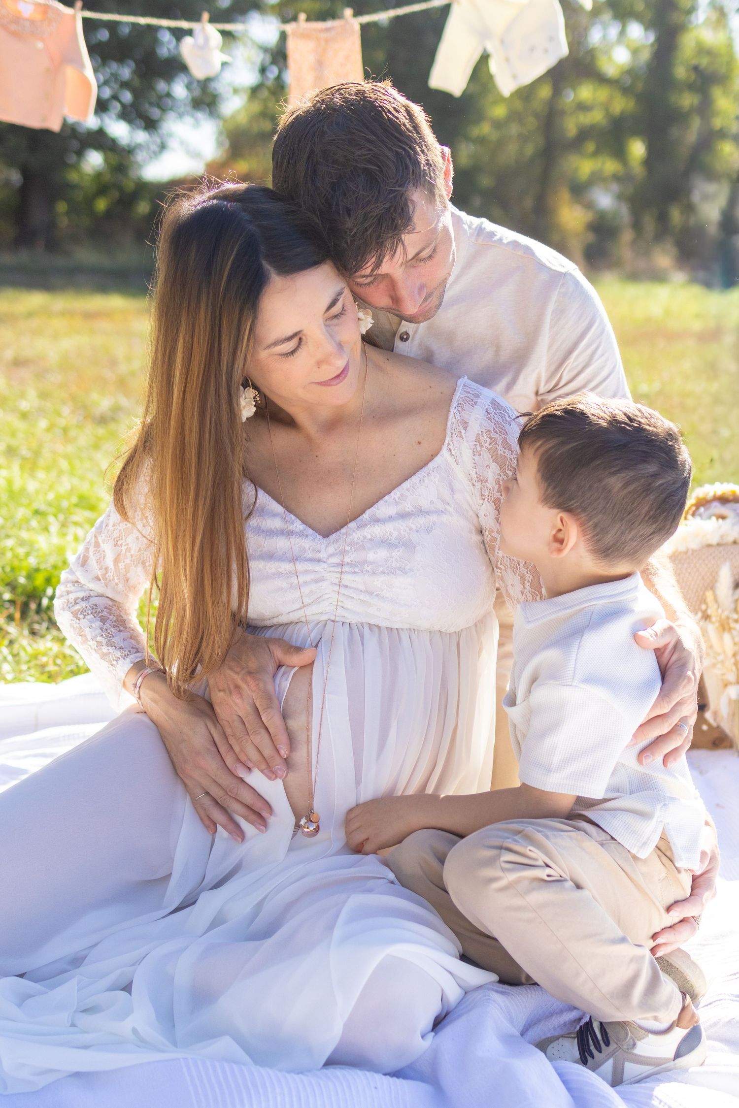 Pregnant woman in white dress sits outdoors with husband and son touching her baby bump during a family maternity photo session.