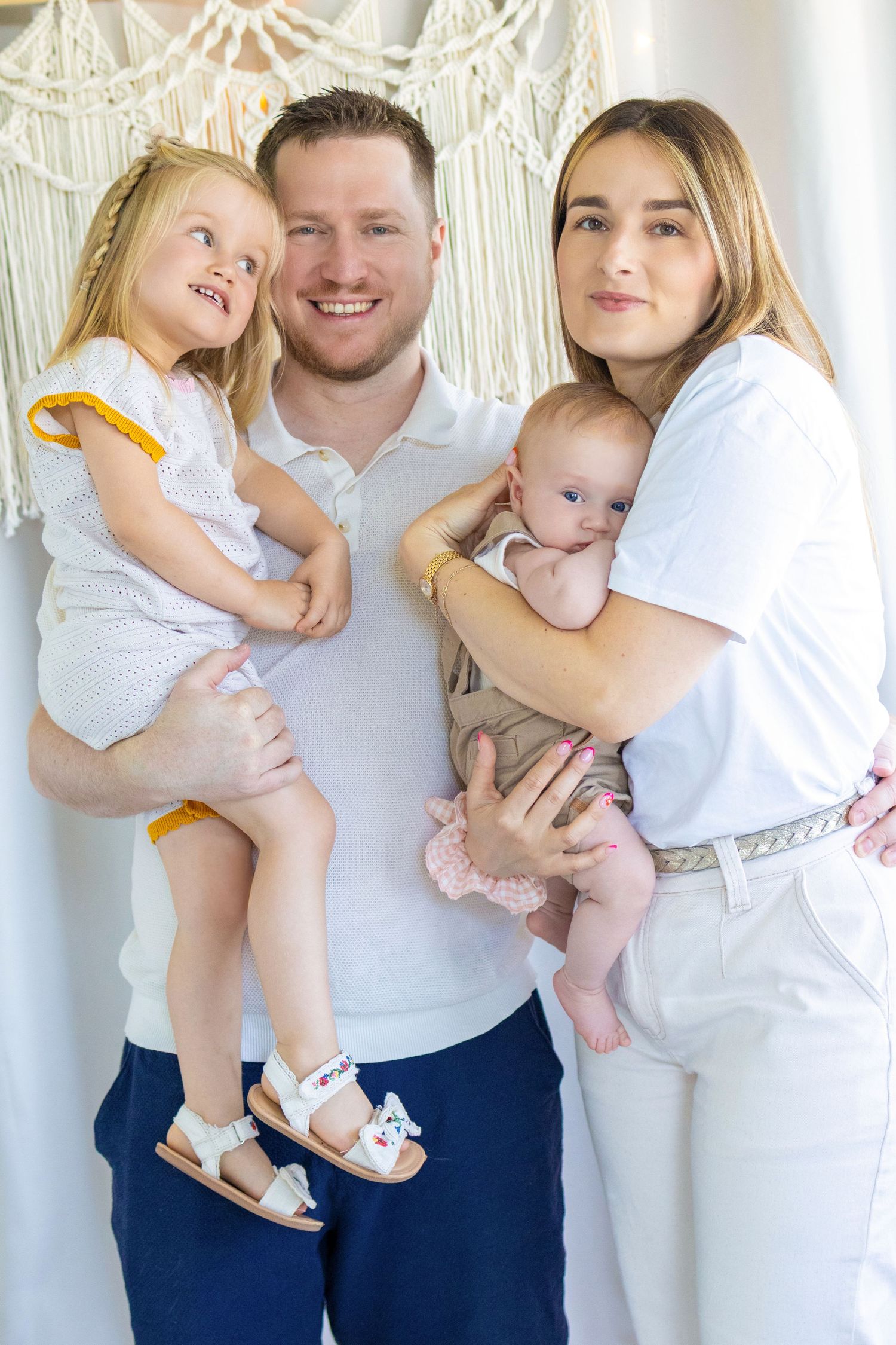Happy family of four posing together, parents holding a toddler girl and newborn baby in a bright studio setting.