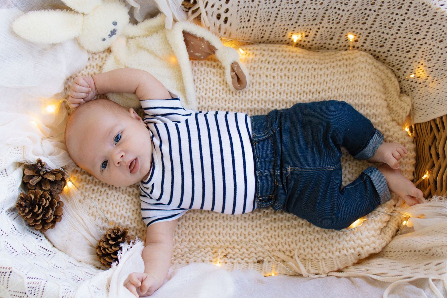 A cute baby in striped onesie and jeans lying in a cozy knit basket with fairy lights and pine cones.