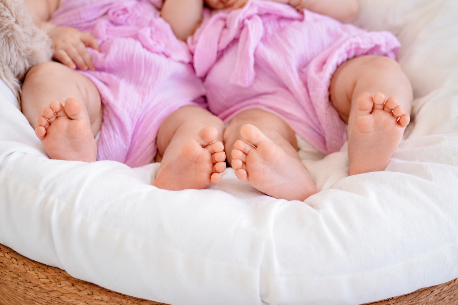 Two newborn twin babies in pink outfits lying on a white pillow, showing their tiny cute feet.