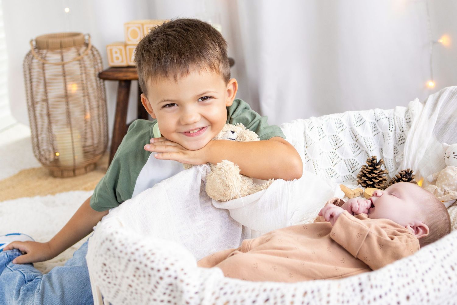 Smiling boy leans over a bassinet holding a stuffed animal next to his sleeping newborn sibling in a cozy nursery.