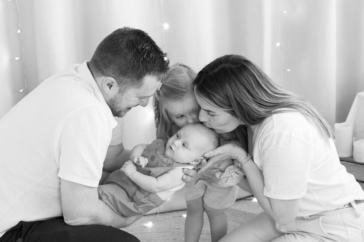 Black and white family photo of parents and young daughter kissing newborn baby together.