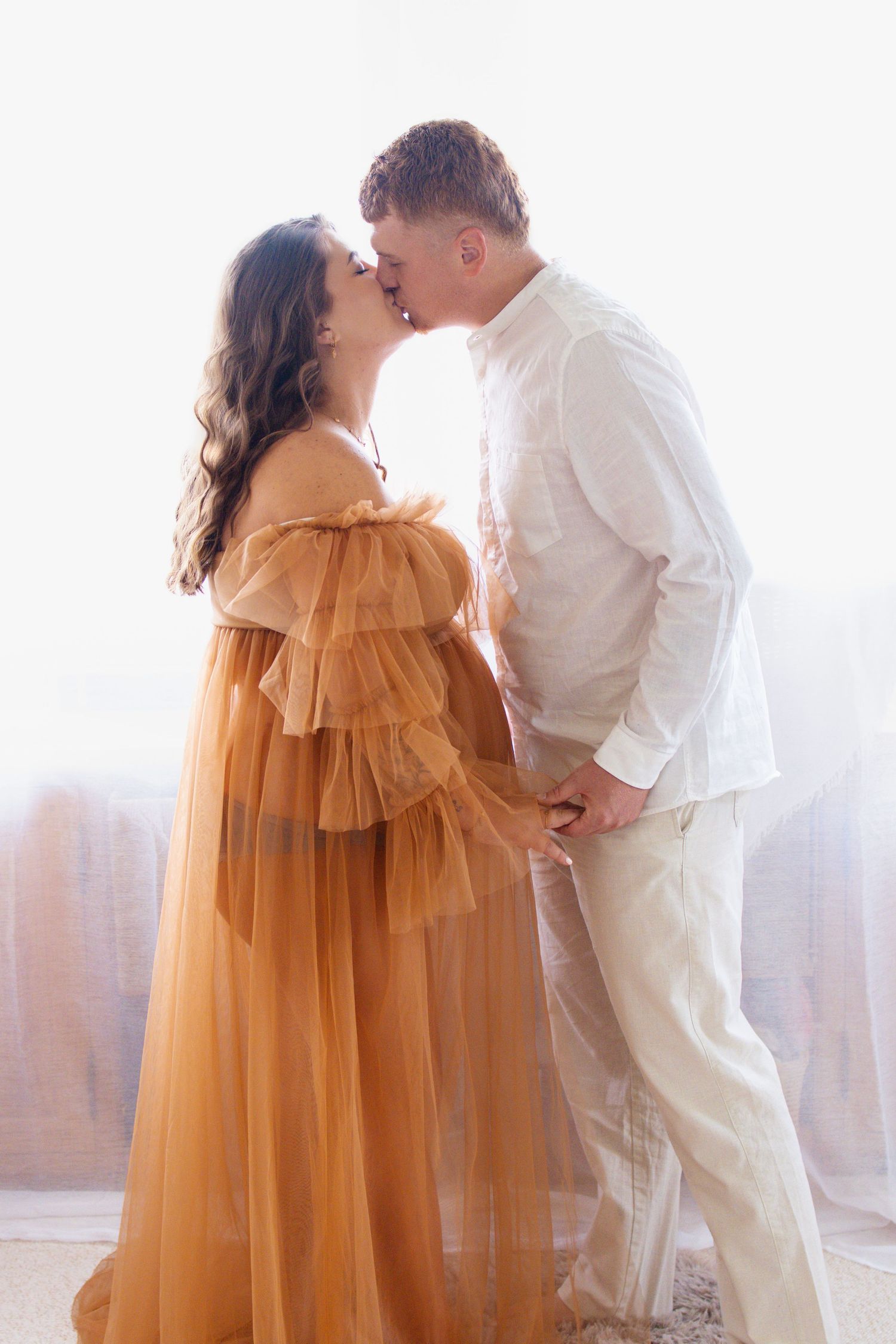 Pregnant woman in golden tulle gown kissing partner in white shirt during romantic maternity photo session.