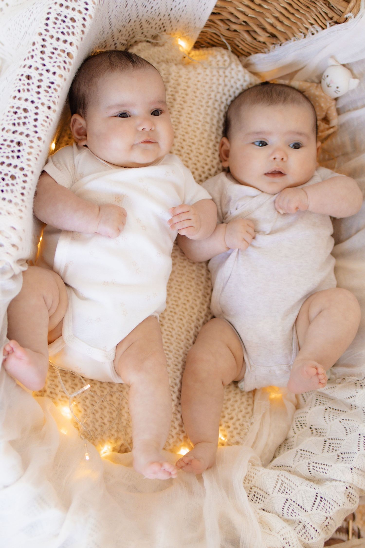 Two adorable twin babies lying side by side in a basket with warm fairy lights, wearing white onesies.