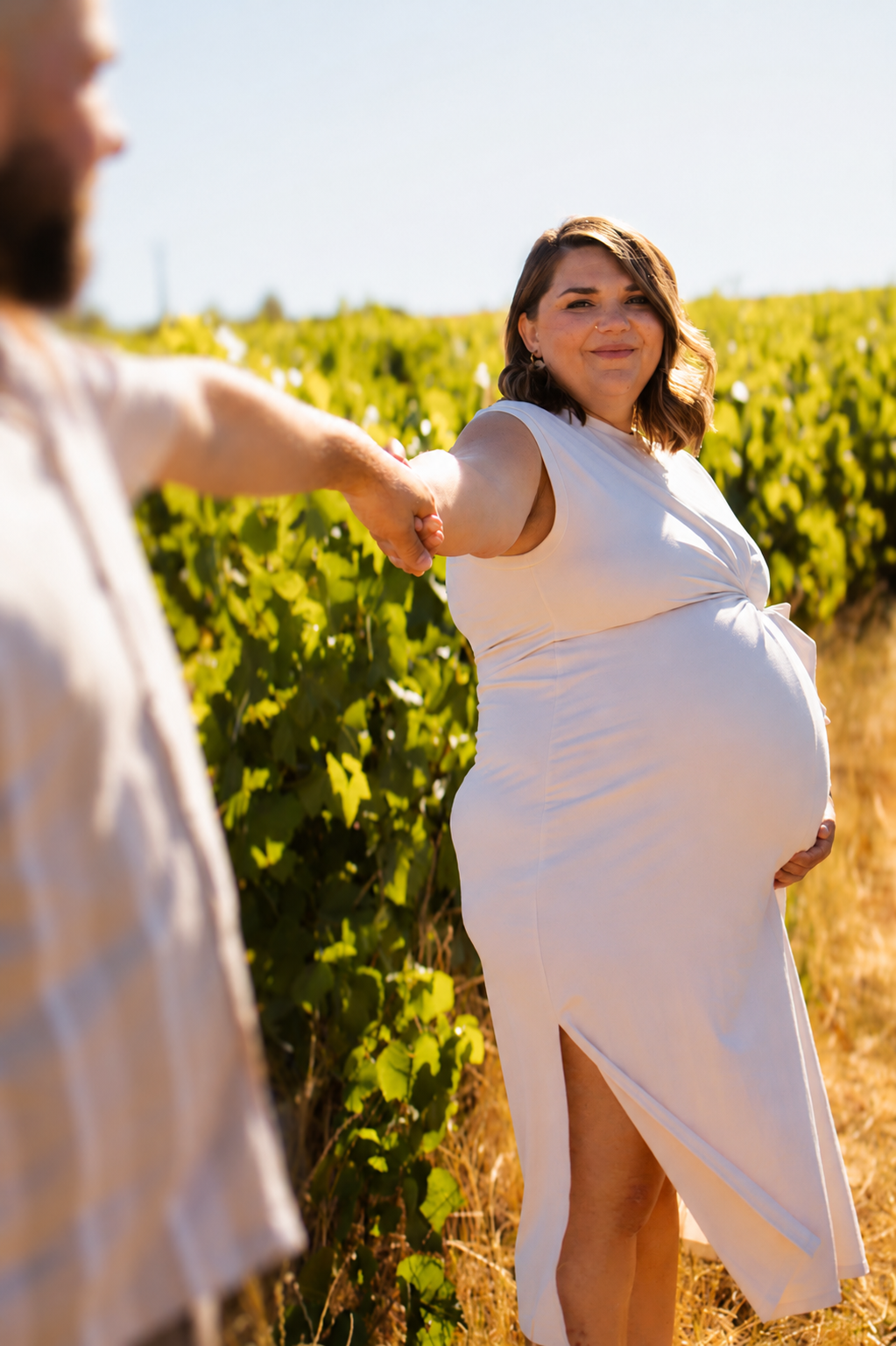 Pregnant woman in white dress holds partner's hand in sunny vineyard, smiling during outdoor maternity photoshoot.