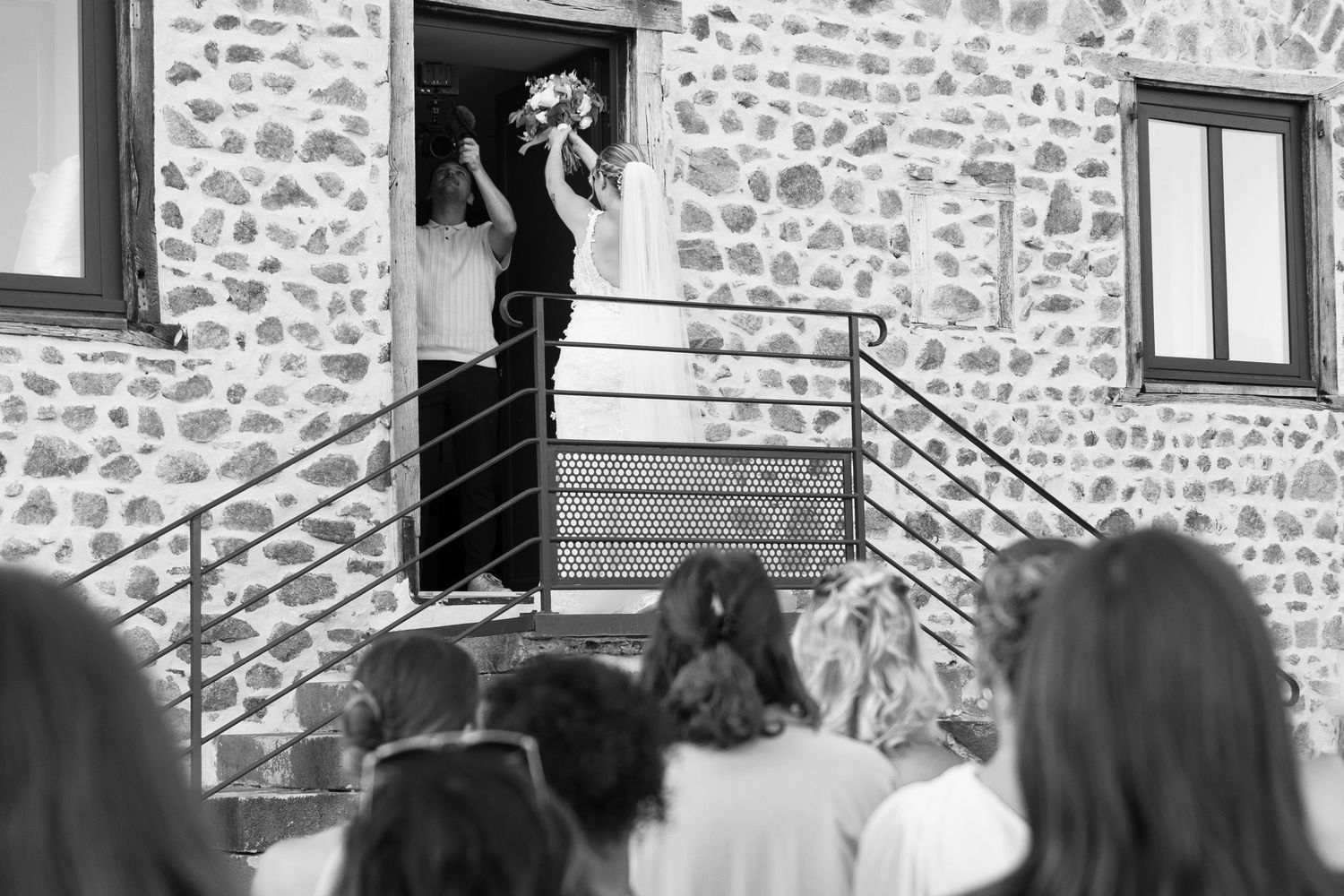 Black and white wedding photo of bride holding bouquet at stone building entrance with guests watching below.
