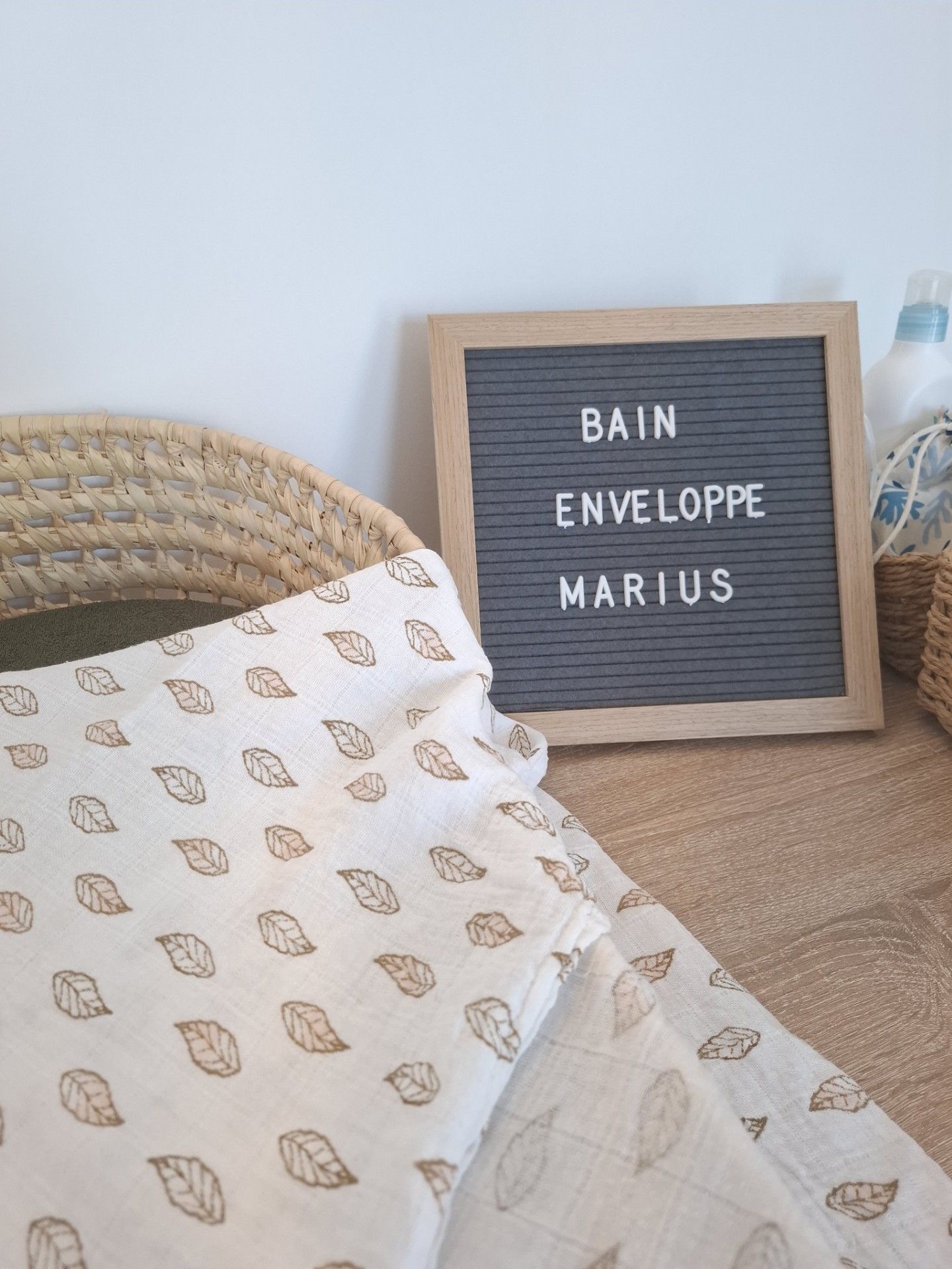 Letter board reading Bain Enveloppe Marius beside a leaf-print muslin cloth and woven basket on a wooden surface.