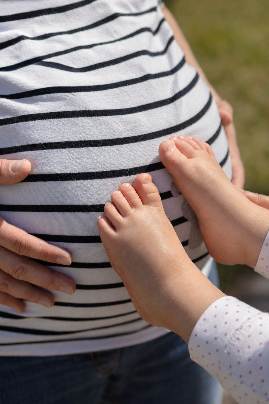 A child's bare feet rest on a pregnant woman's belly in a striped shirt outdoors.