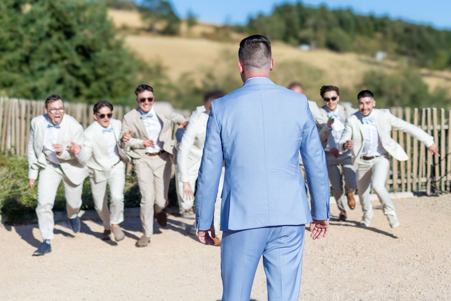 Groom in blue suit faces excited groomsmen in tan suits celebrating outdoors at a sunny wedding venue.