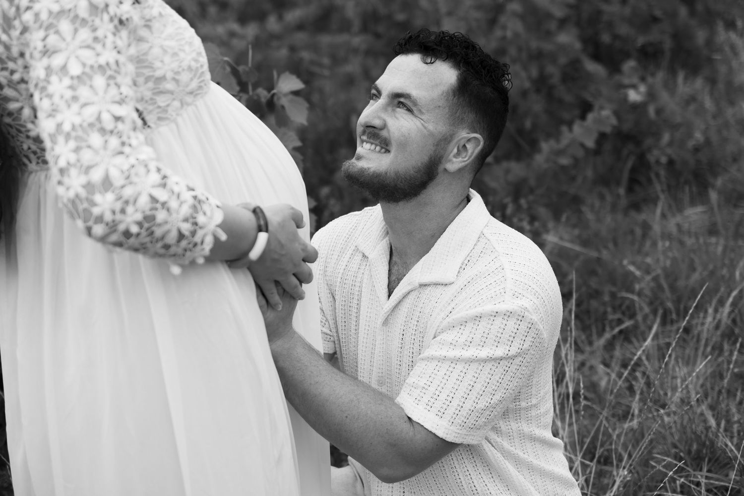 Black and white photo of a man kneeling before a pregnant woman, lovingly holding her baby bump outdoors.