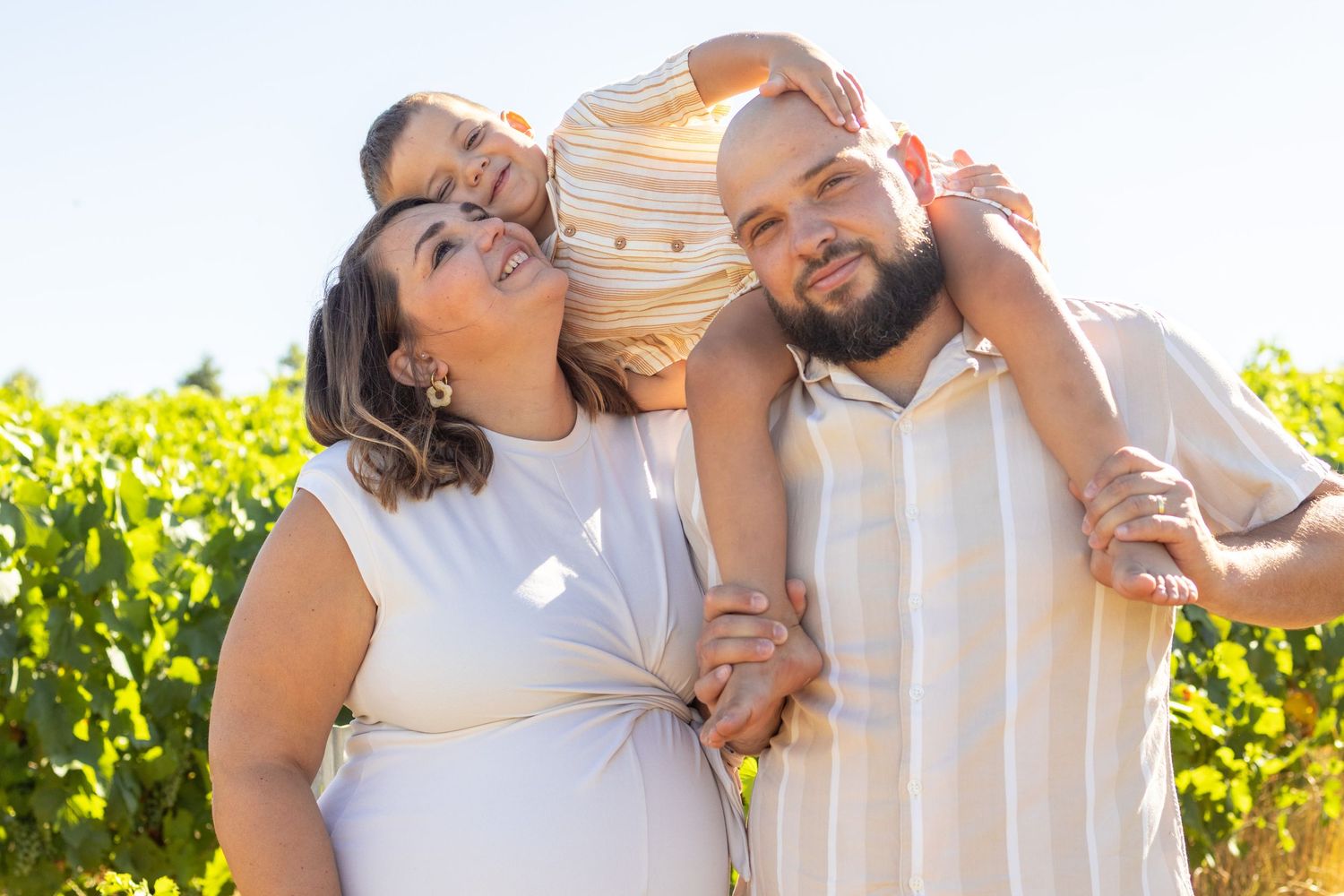 Happy family of three posing outdoors in a sunny vineyard, parents smiling with child on shoulders.