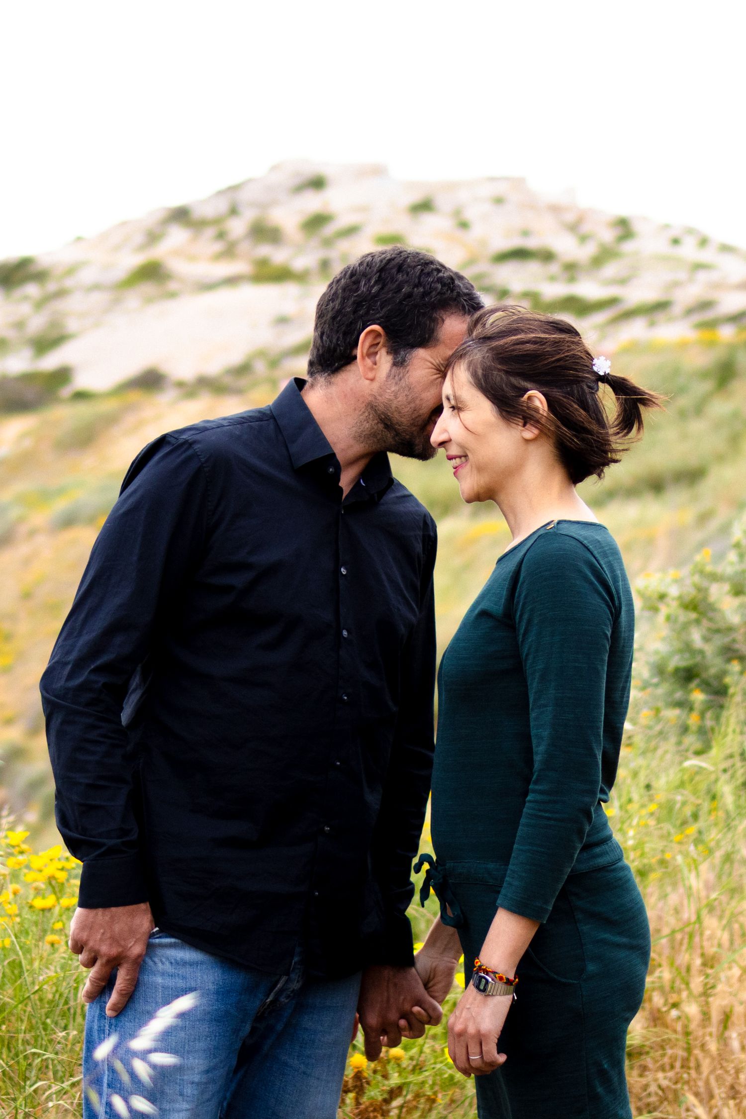 Romantic couple holding hands in a wildflower field, man in black shirt leaning toward smiling woman in green dress.